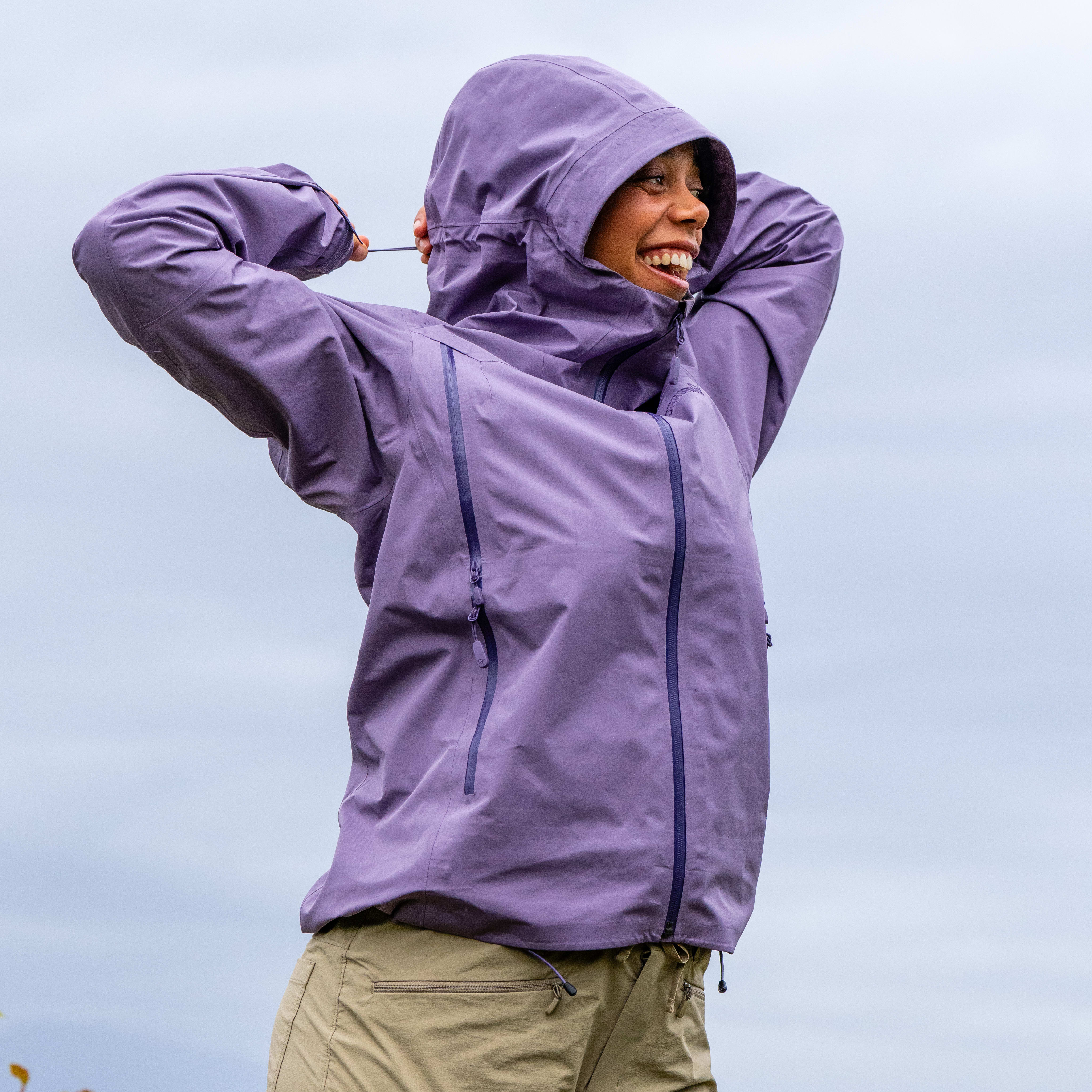 Smiling woman wearing a purple Norrøna hooded jacket and beige Norrøna pants outdoors.