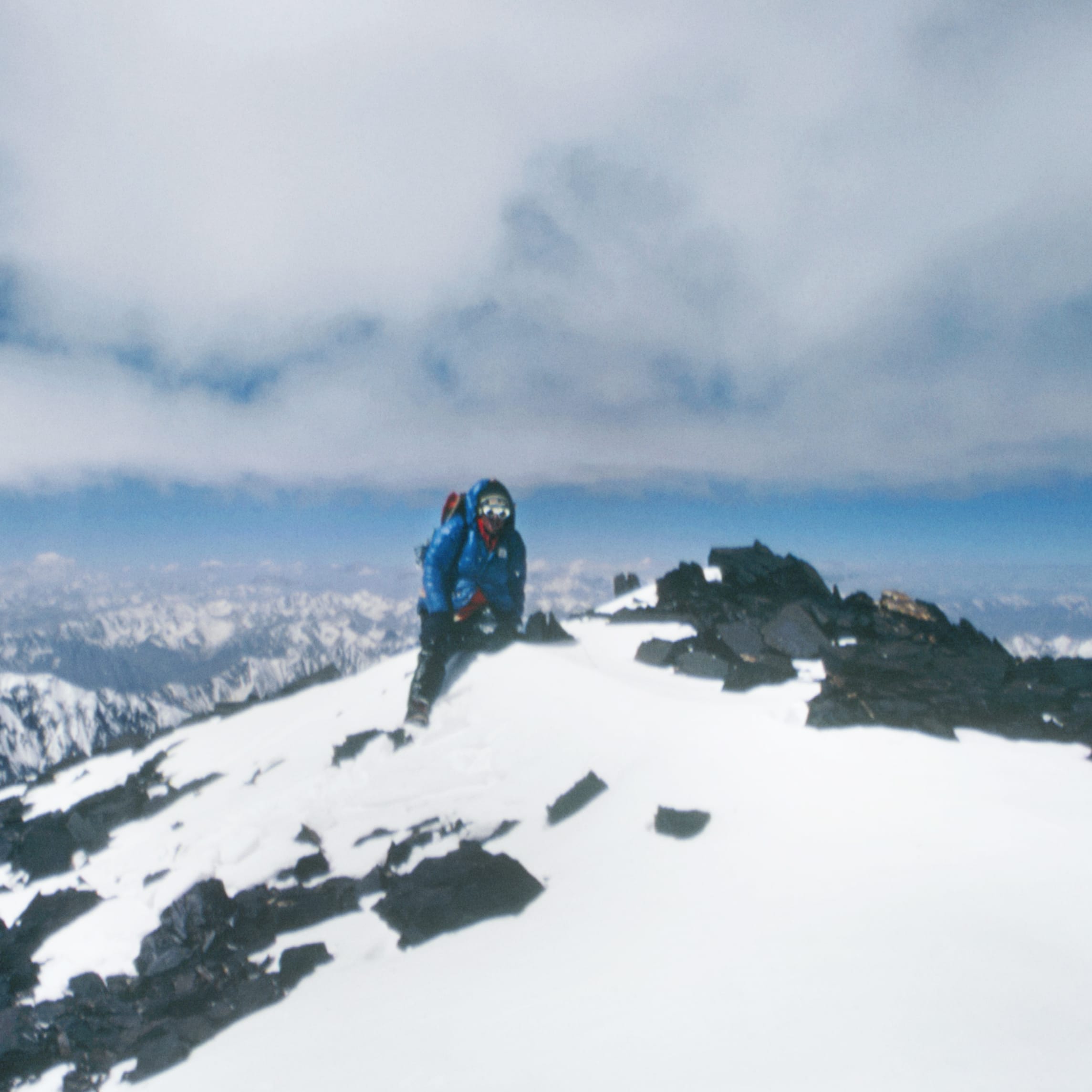 A person in a blue jacket and a backpack stands on the summit of a snowy mountain. A range of mountains is in the background. The sky is blue with clouds.
