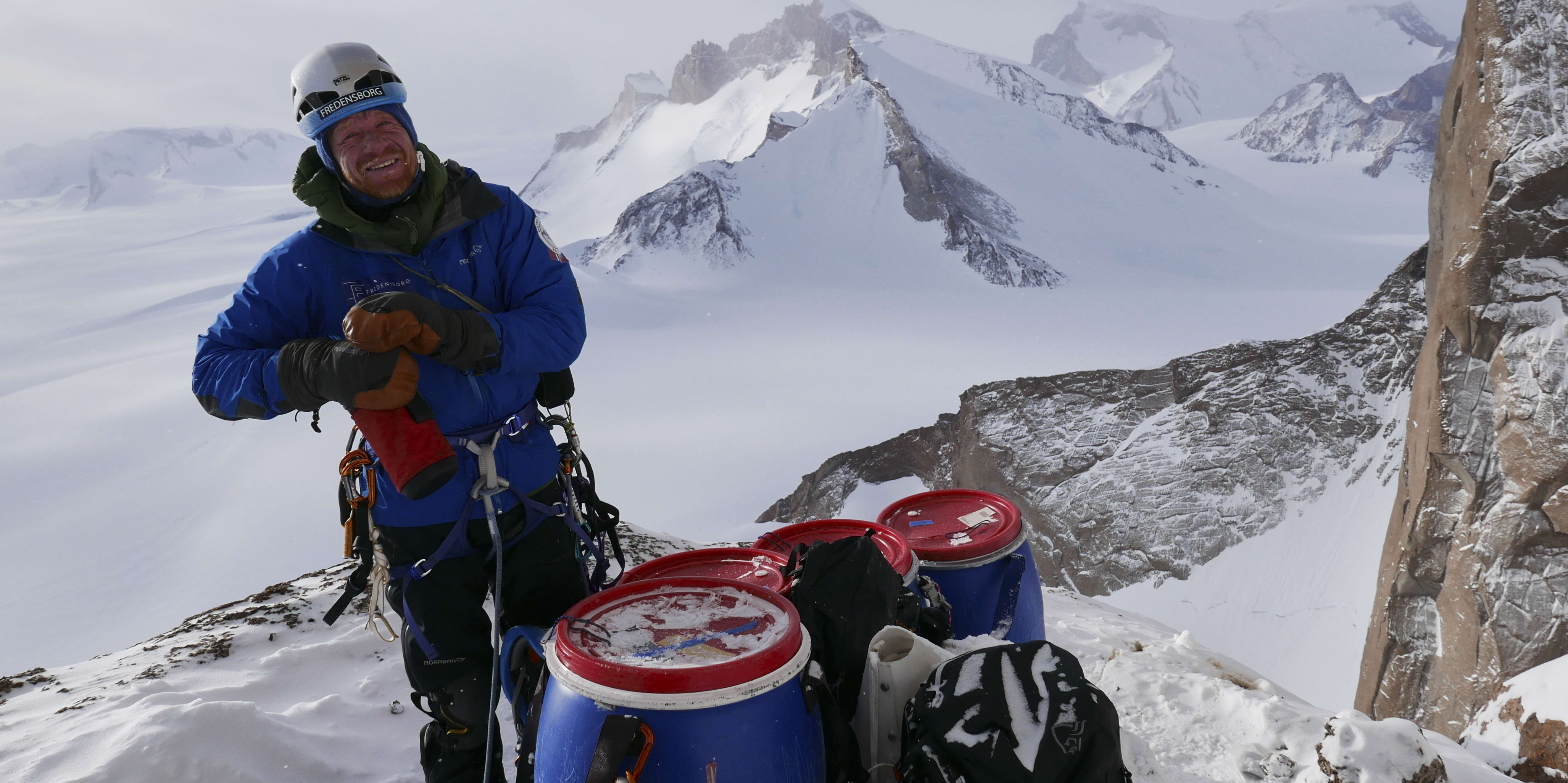 A man wearing a Norrøna jacket and helmet smiles on a snowy mountain peak. He is standing near climbing gear and a large, red and blue barrel.