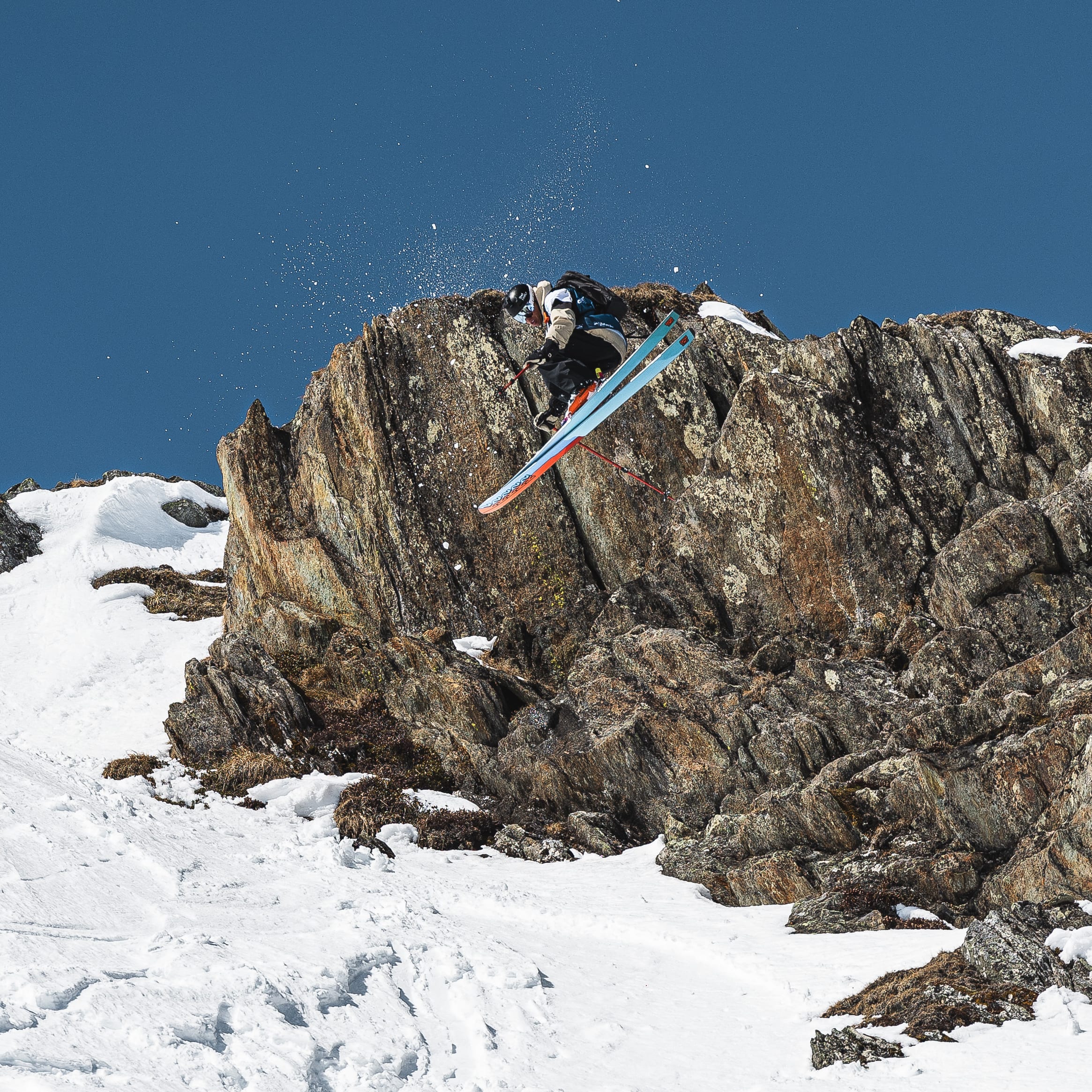 A skier wearing Norrøna clothing is airborne off a rocky cliff face on a sunny, snowy mountain. Another skier is visible in the background.