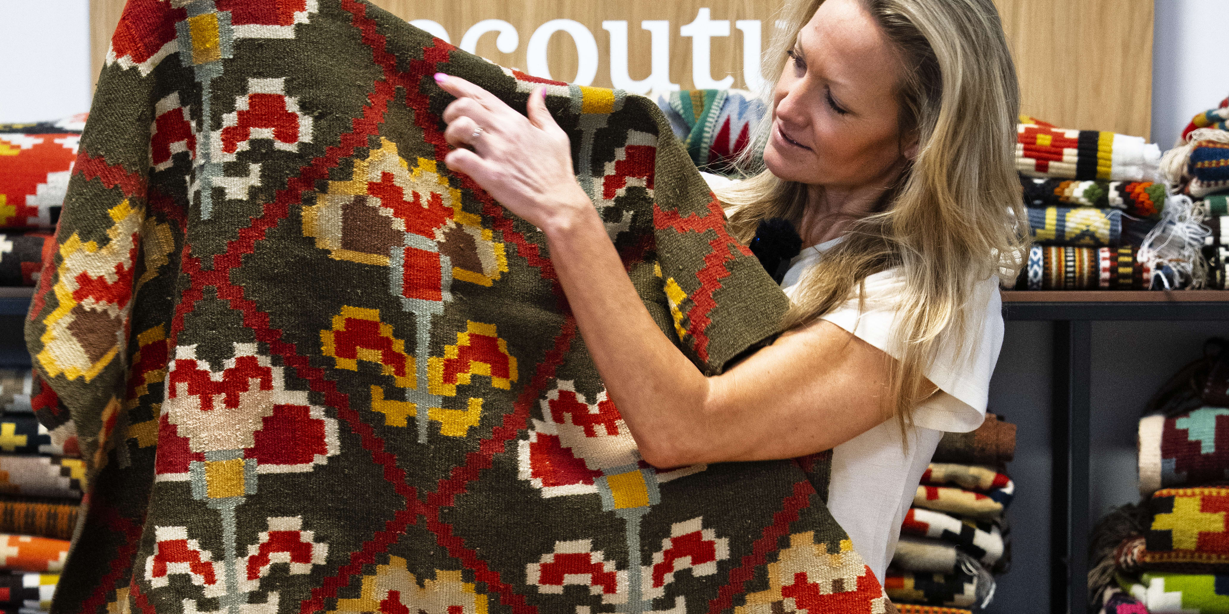 A woman holds up a patterned rug in a shop with other rugs. The rug has a green background with red, yellow, and brown floral patterns.