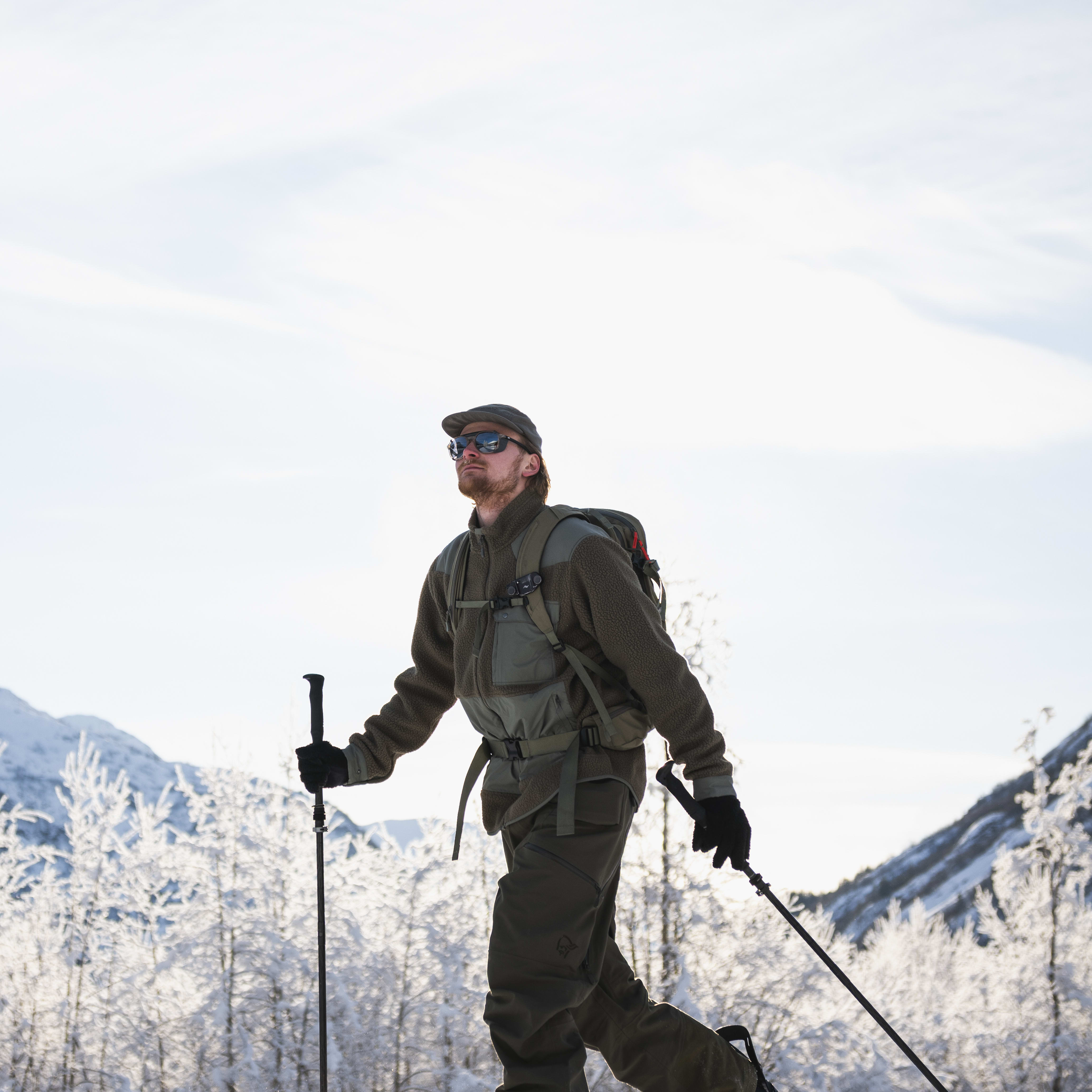 A man snowshoes, wearing Norrøna jacket and pants, backpack, hat and sunglasses, using poles against a snowy mountain backdrop.