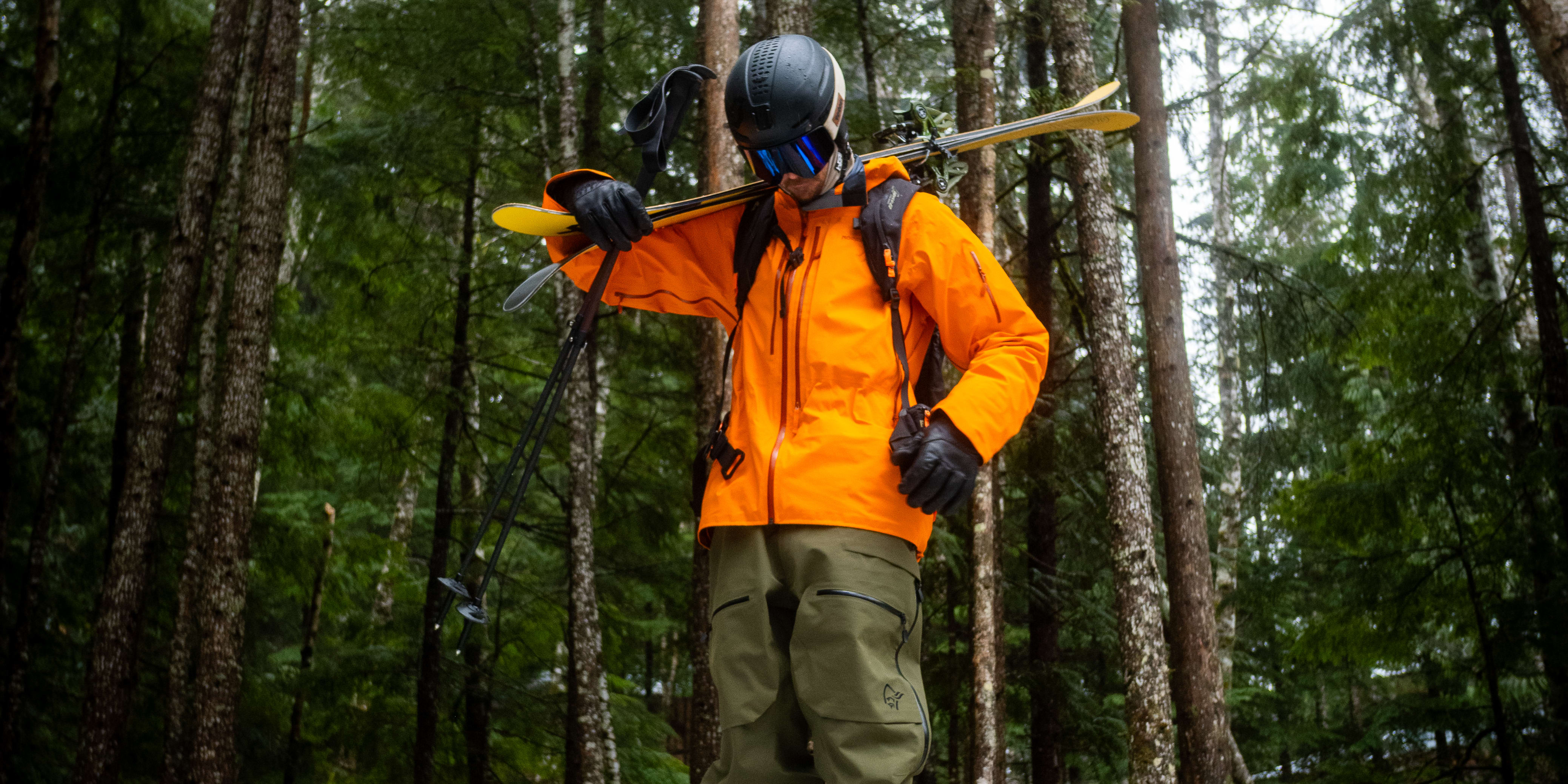 A skier stands on a stump in a forest, skis over shoulder. They wear a helmet, goggles, and a Norrøna orange jacket and green pants.