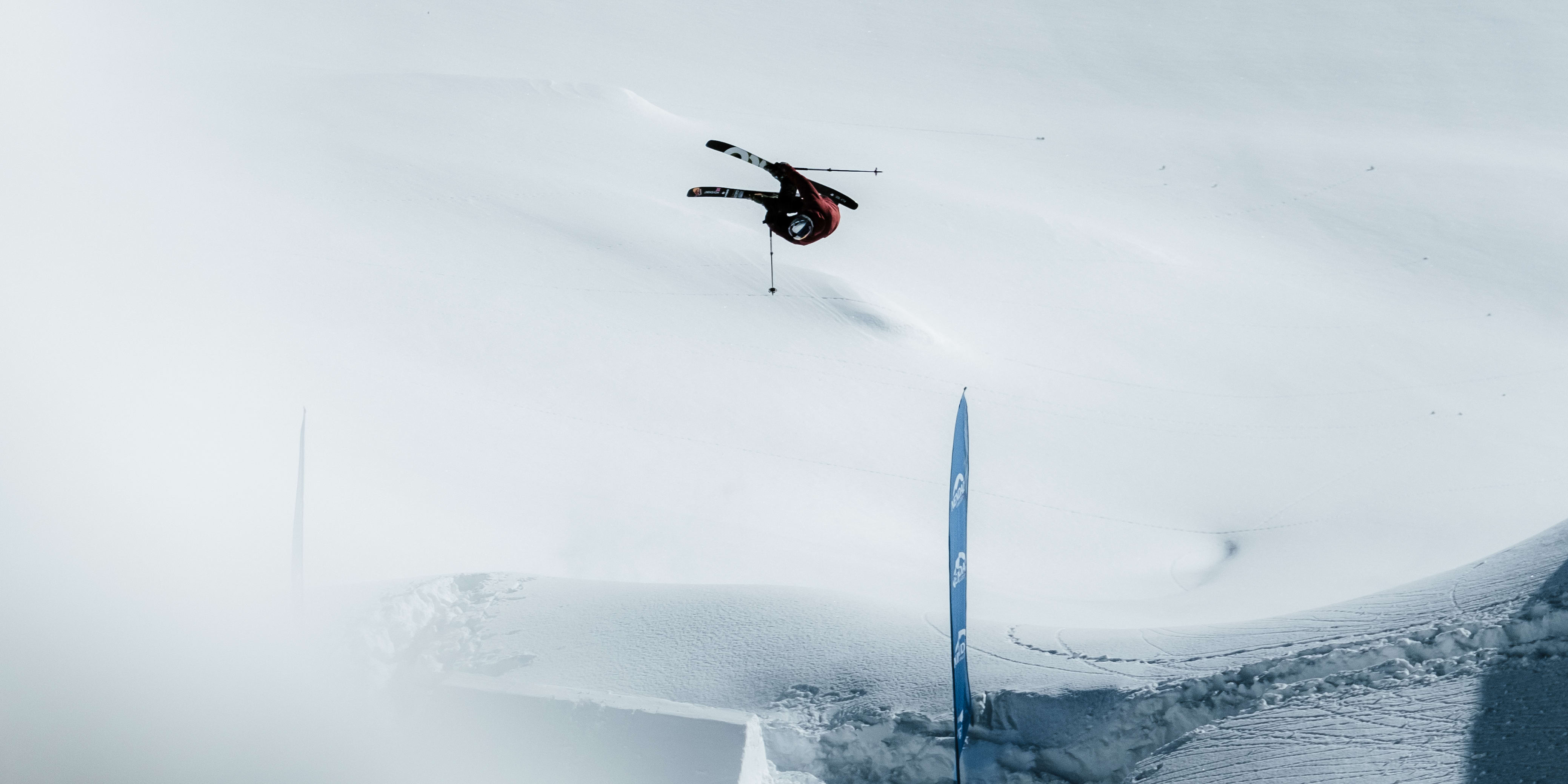 A skier wearing Norrøna clothing performs a trick mid-air over a snowy jump, a blue flag marker to the right.

