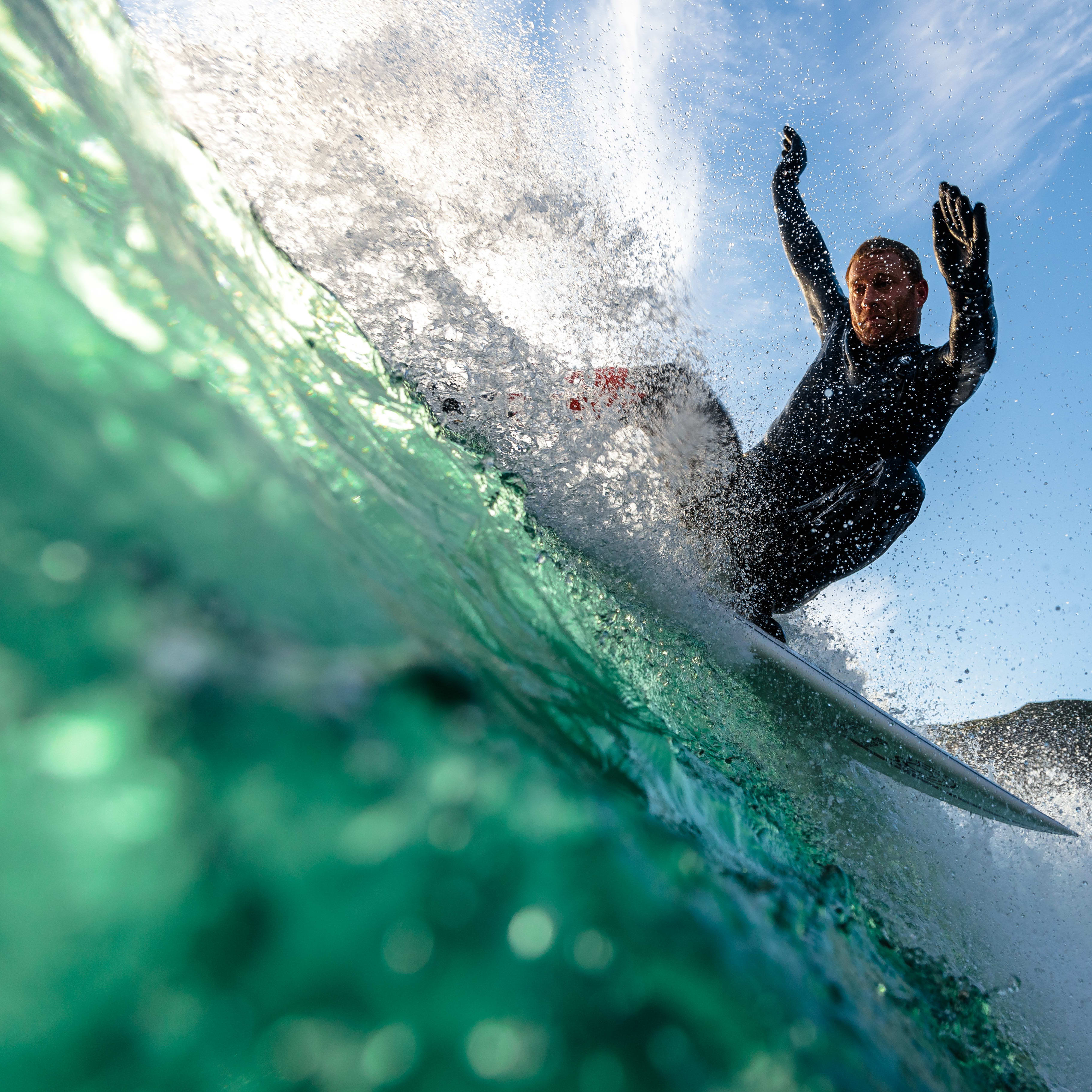 A surfer in a Norrøna wetsuit rides a wave, arms raised, with splashing water and a blue sky visible.