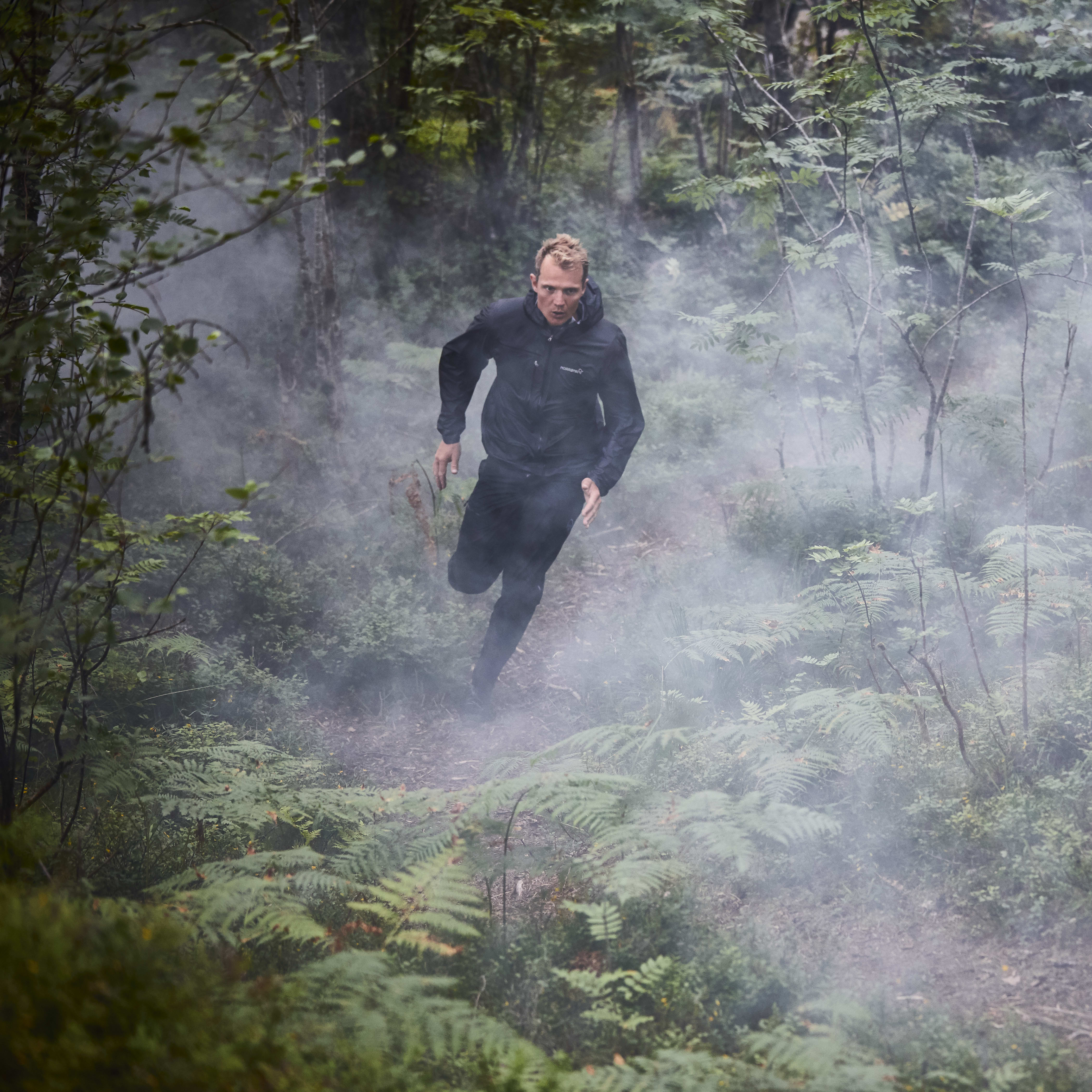 A man wearing a Norrøna jacket runs through a foggy forest.  
