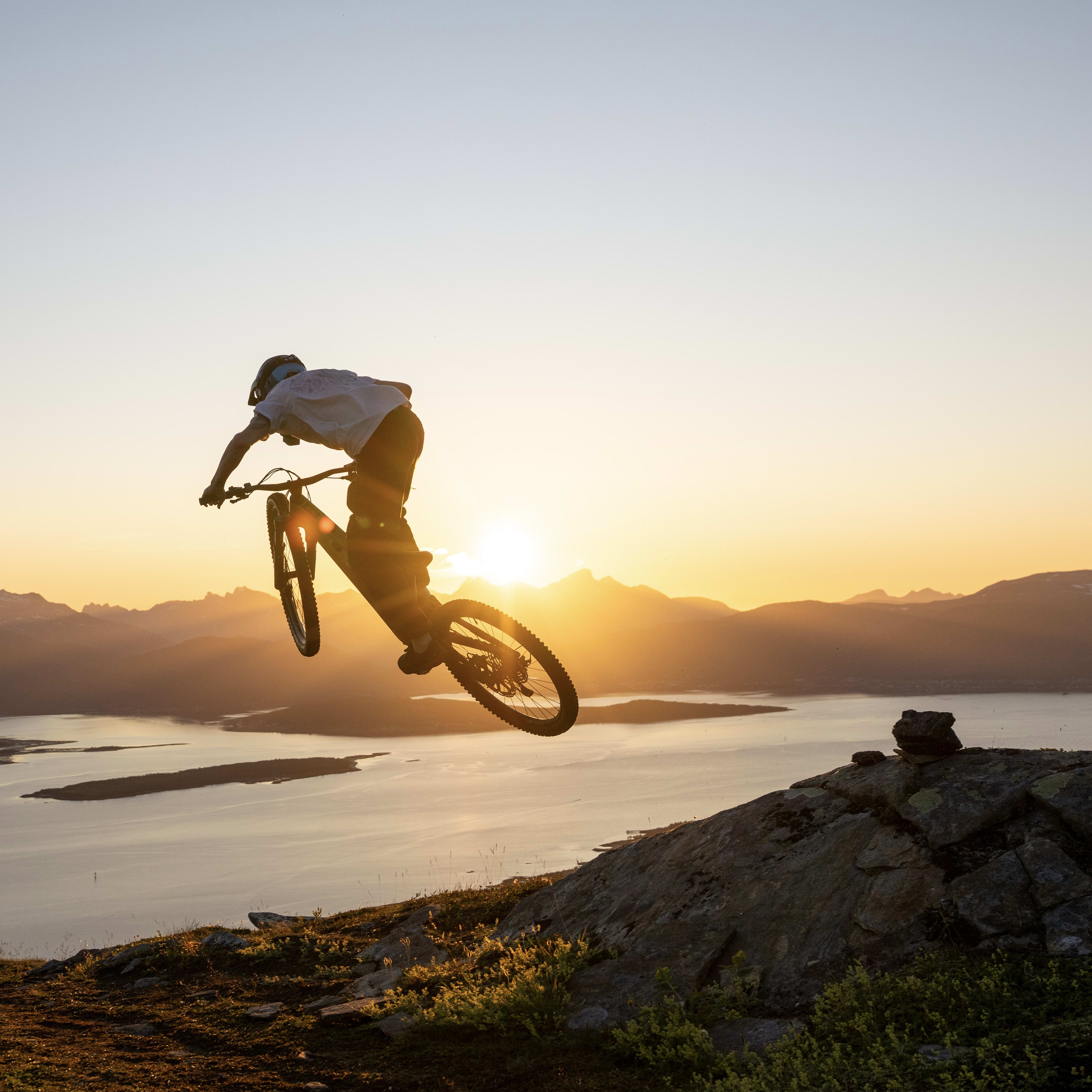 A person on a mountain bike jumps, silhouetted against a sunset with mountains in the distance. They are wearing a helmet and a Norrøna shirt.