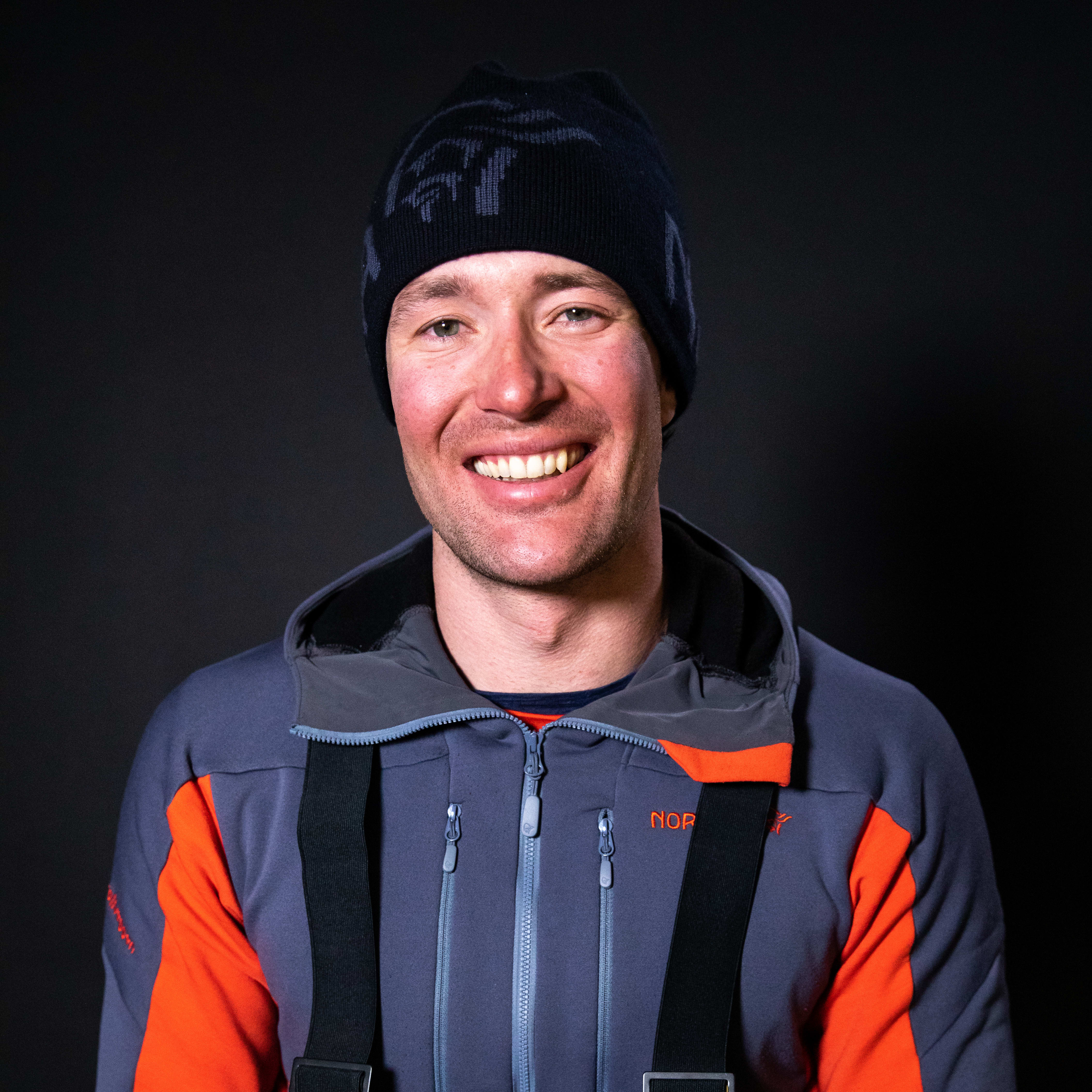 Smiling man in a Norrøna beanie and grey/orange jacket with suspenders against a dark background.