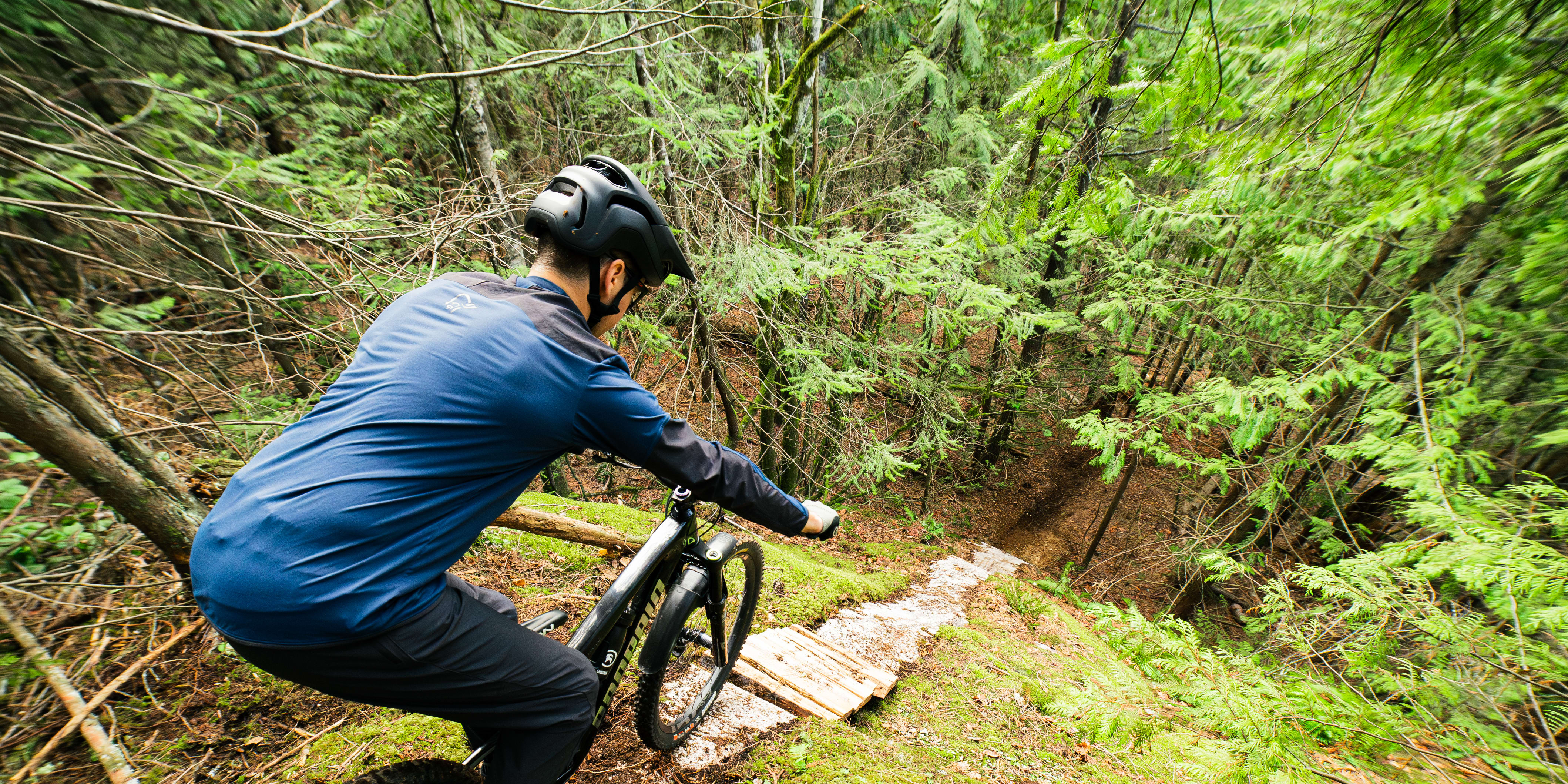 A cyclist wearing a helmet and a blue Norrøna jacket rides a mountain bike on a trail through a lush green forest.