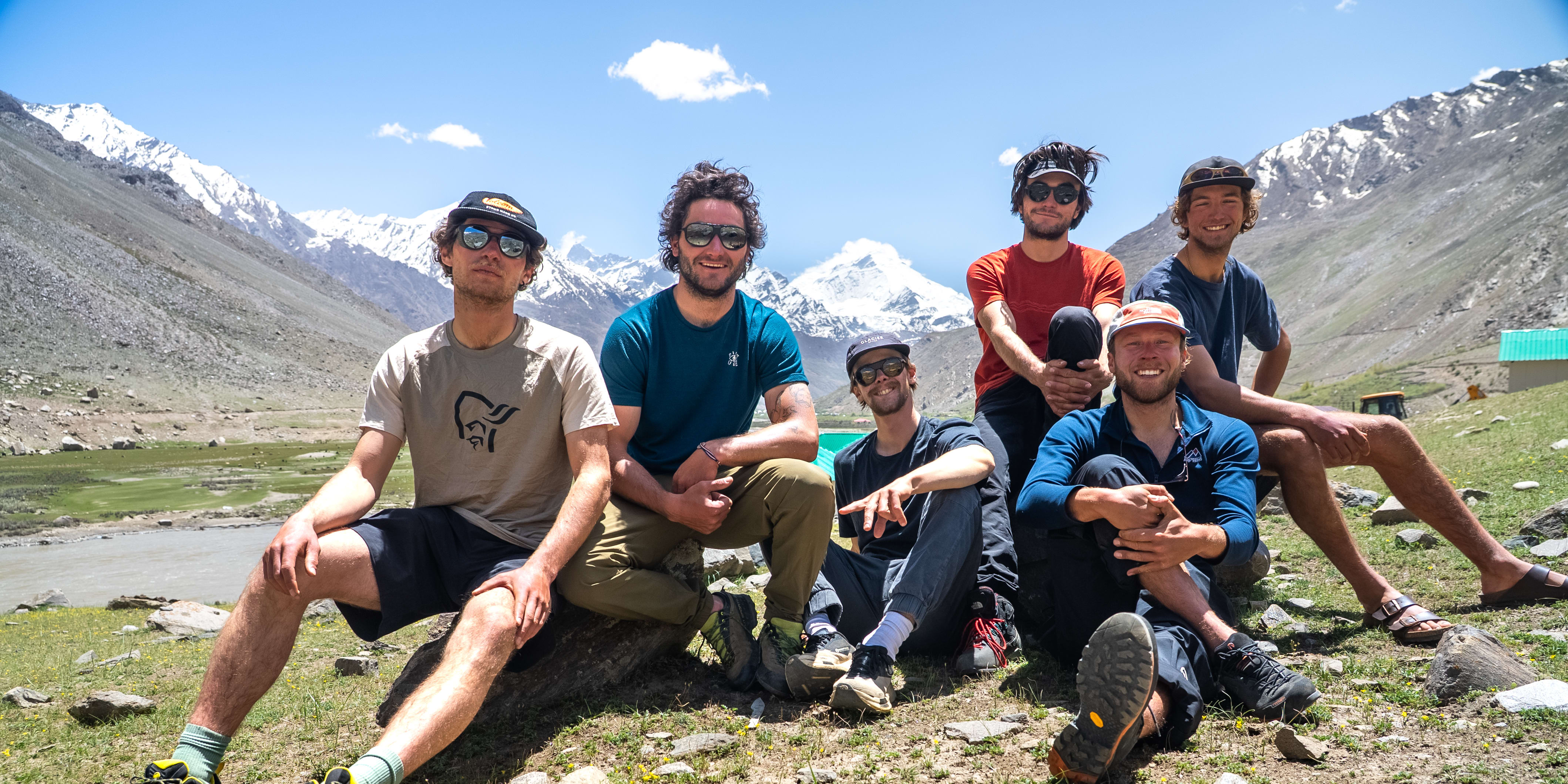 Six smiling people, one in a Norrøna shirt, pose with snow-capped mountains and a river in the background on a sunny day.