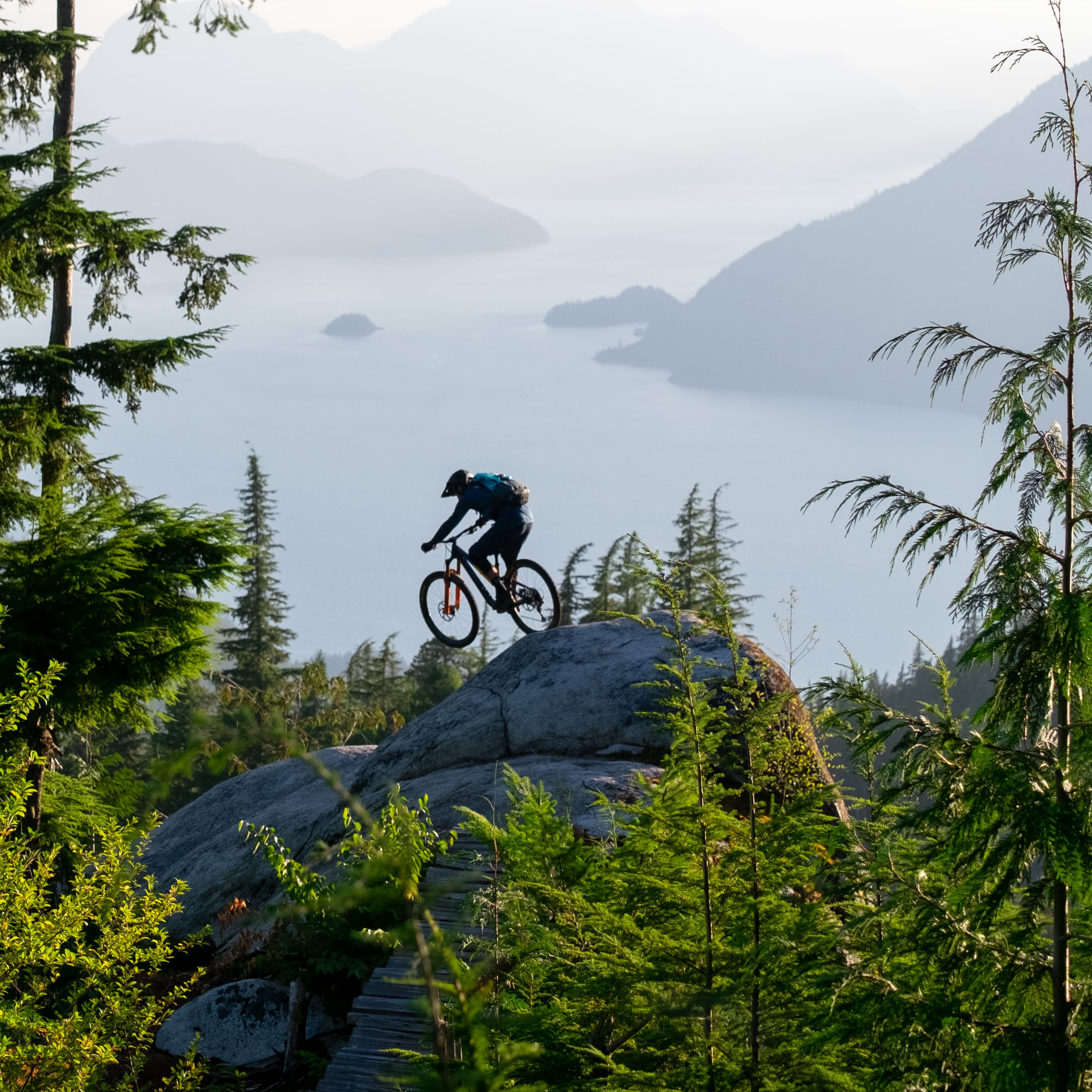 A mountain biker wearing Norrøna clothing rides on a rock outcrop. A lake with distant mountains forms the backdrop.