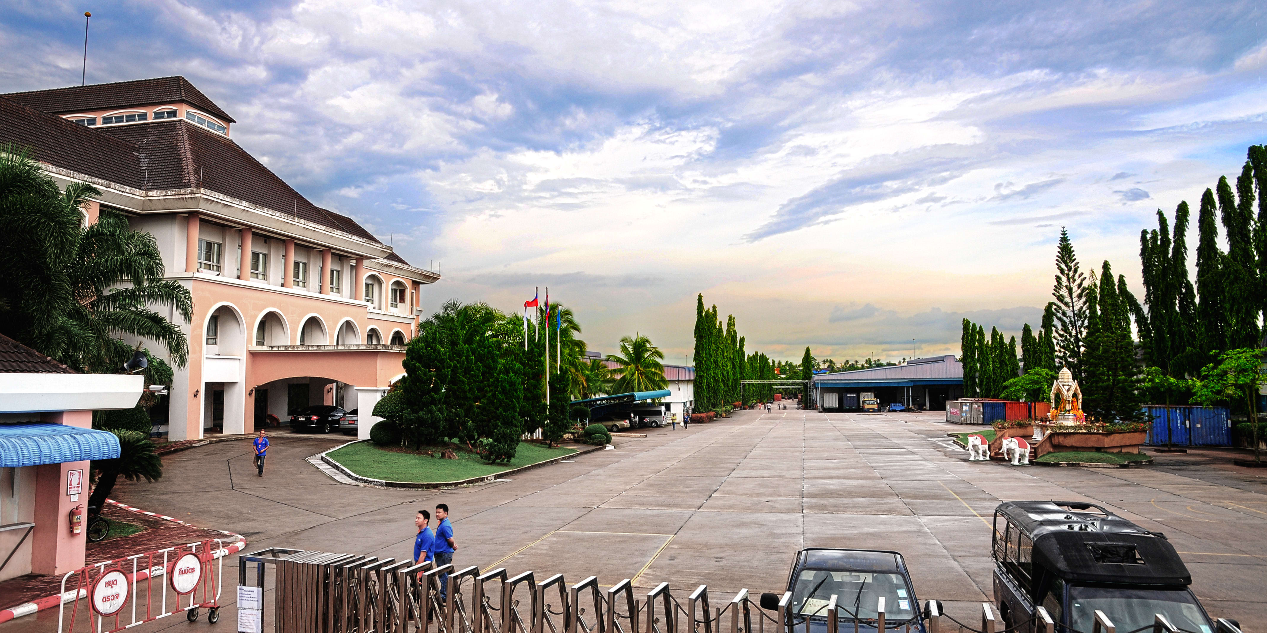A large industrial complex with an open parking lot, a large pink building in the foreground, and a large concrete building with a large metal roof in the background. There are two men in blue shirts walking on the paved parking lot, and there is a vehicle in the background with its trunk open.