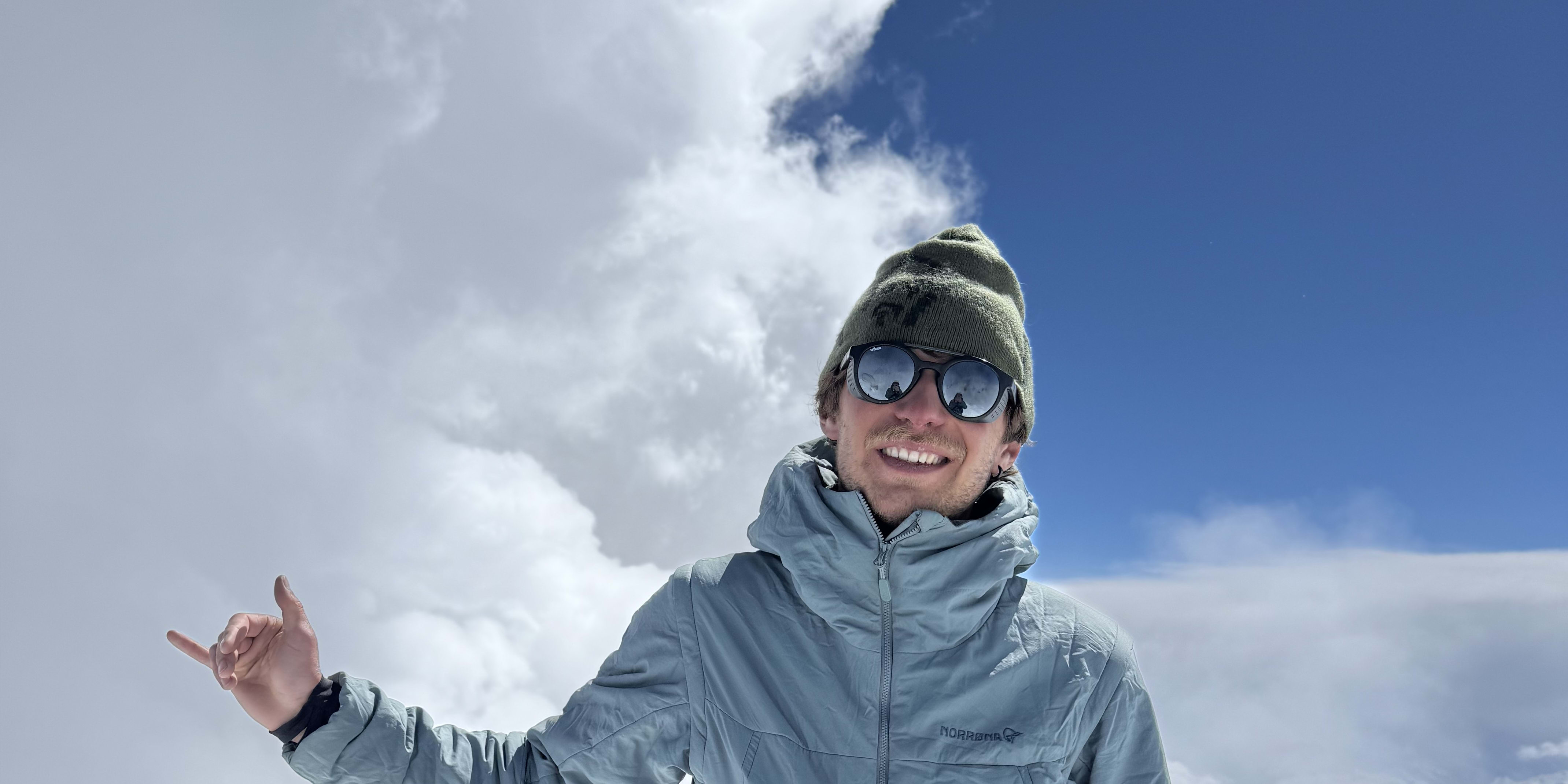 A climber smiles in a Norrøna jacket, harness, and hat, on a snowy peak with a bright blue sky and clouds. They gesture with a finger point. A backpack rests behind.