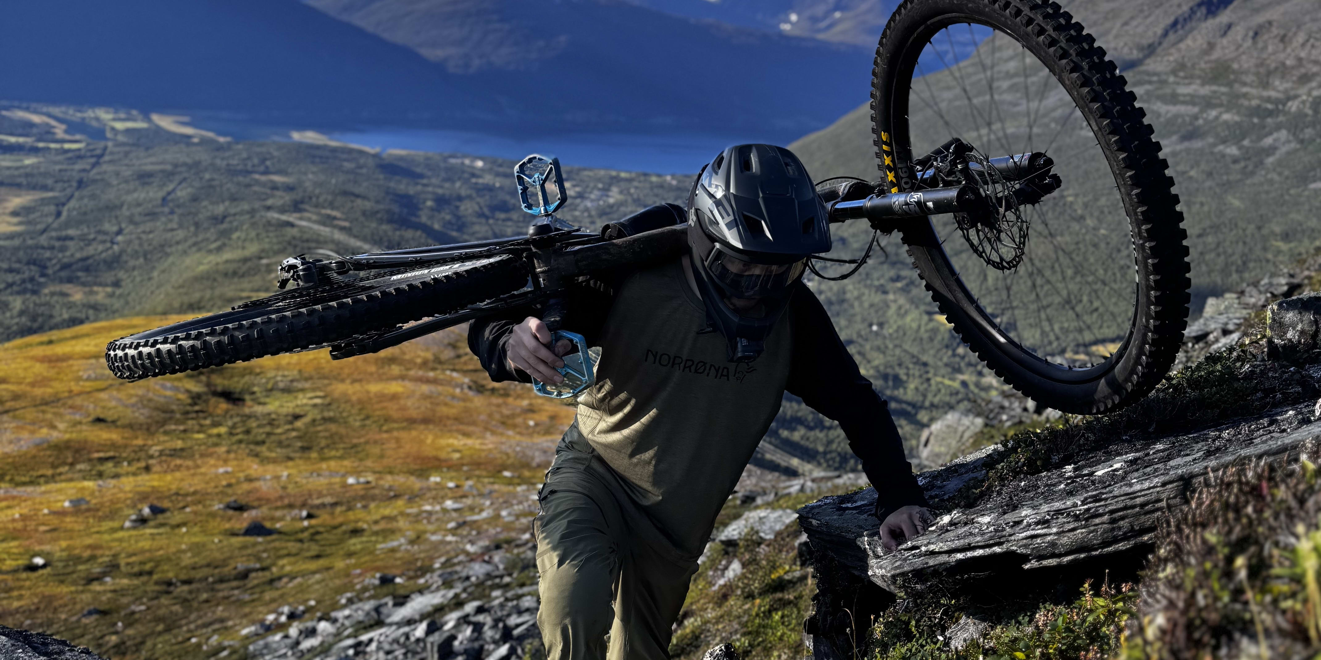 A cyclist in Norrøna clothing carries their bike up a rocky hill. Mountains and blue sky are visible in the background.

