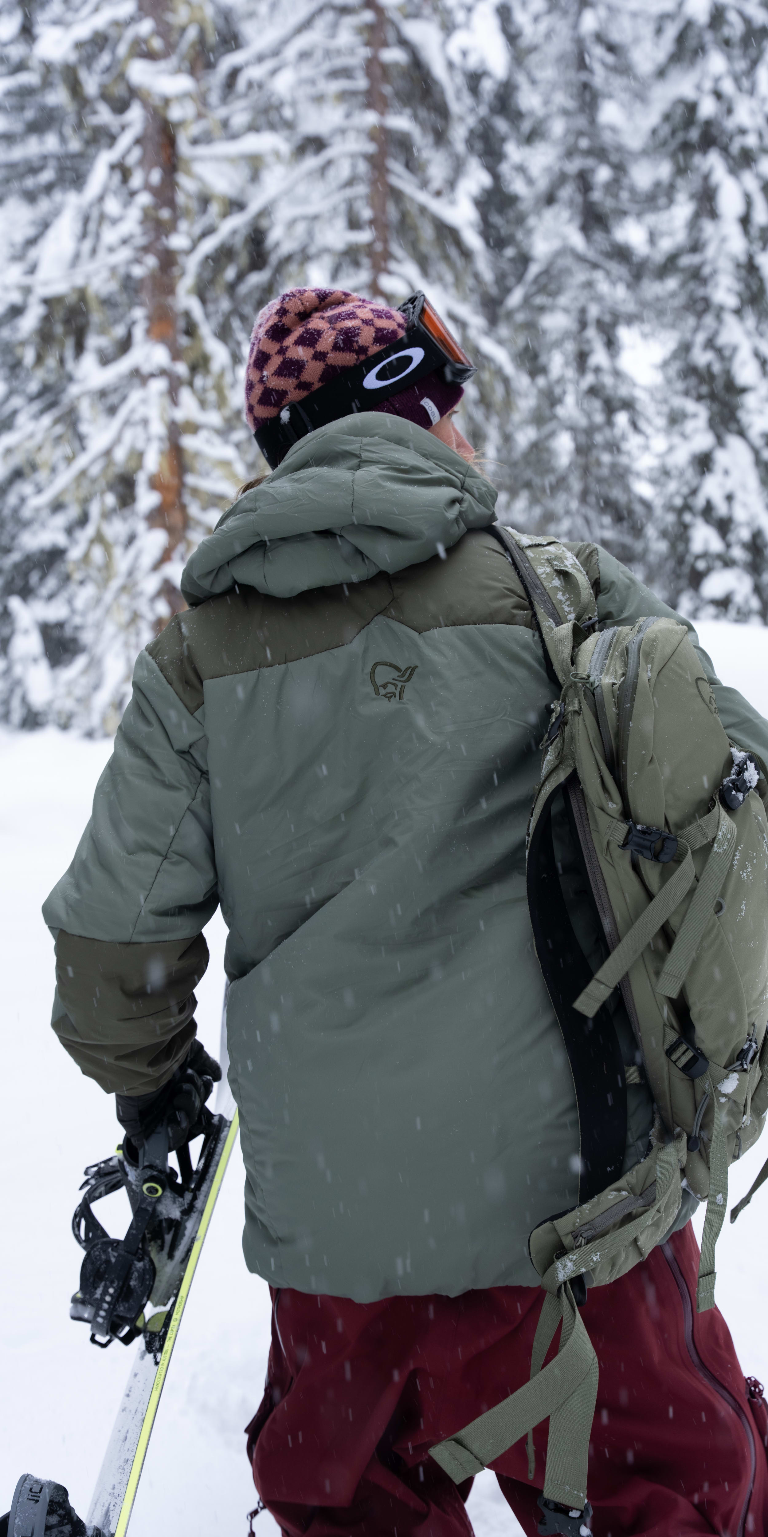 Back view of a person with a snowboard in a snowy forest. They wear a Norrøna jacket and backpack.
