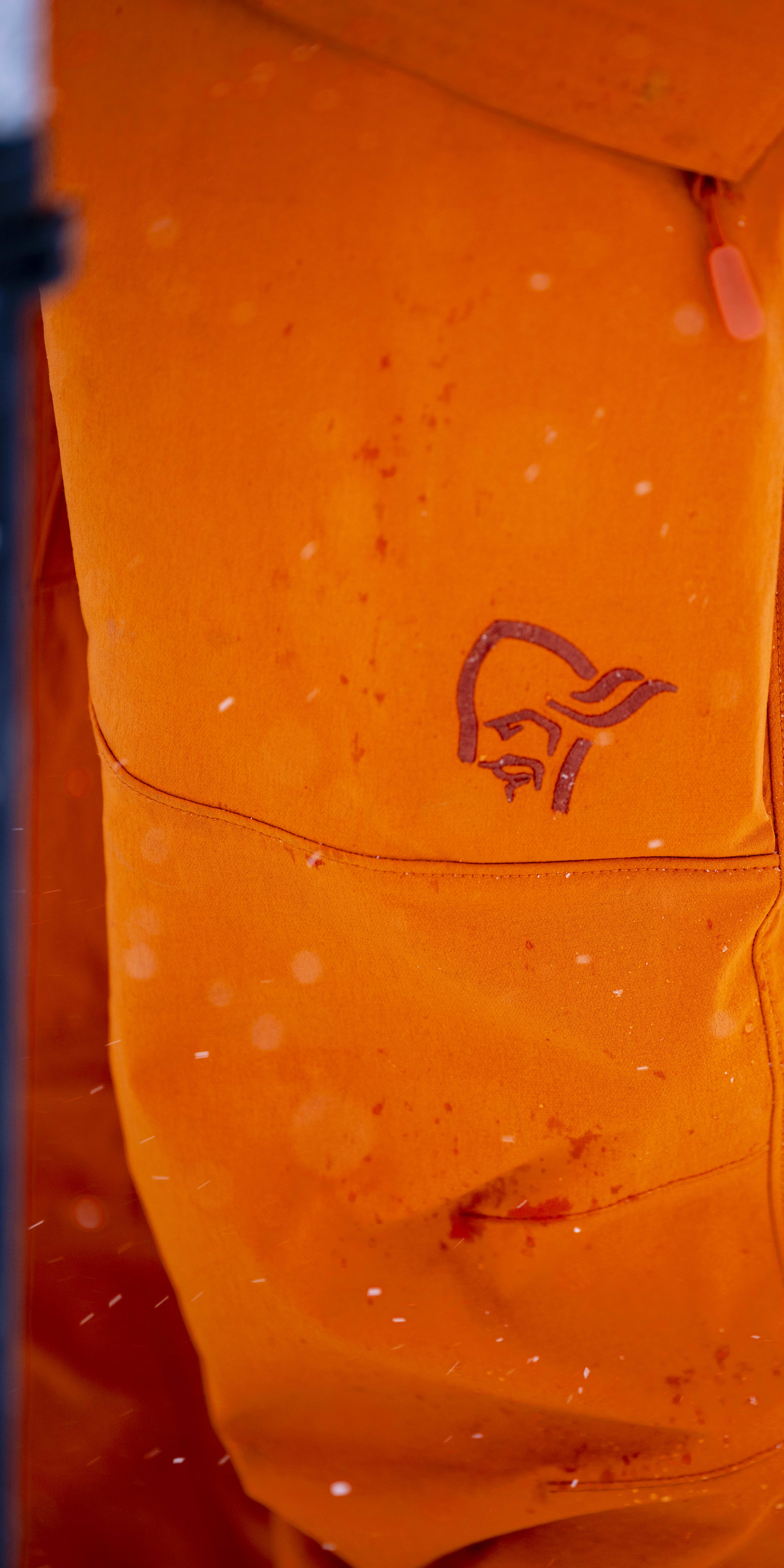 A close up of the right leg of an orange Norrøna  ski pant. The pant has a zipper on the side and a Norrøna logo above the knee.
