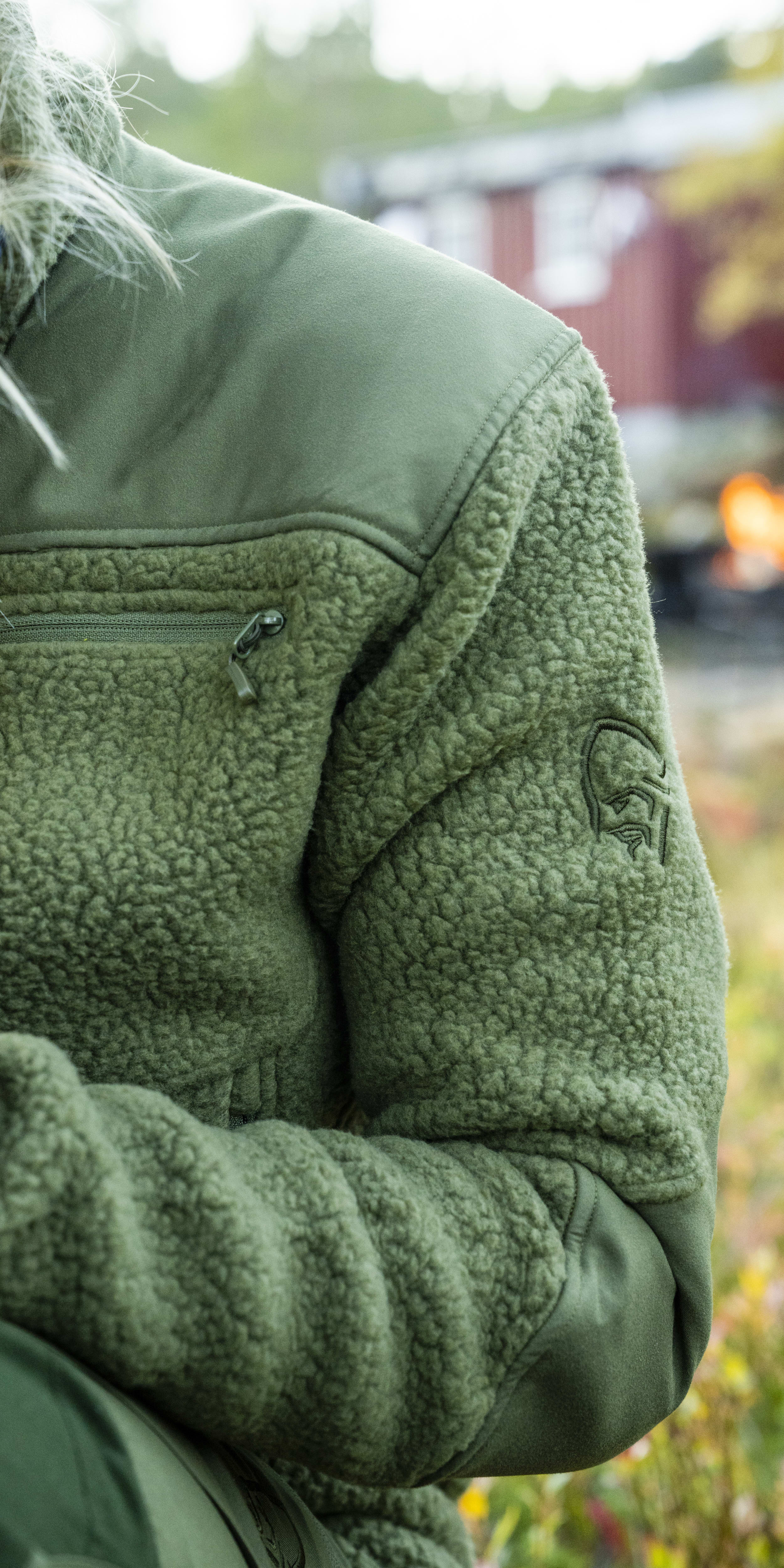 Close up of a green Norrøna fleece jacket with a zippered pocket.  The jacket is in front of a blurry background of greenery.