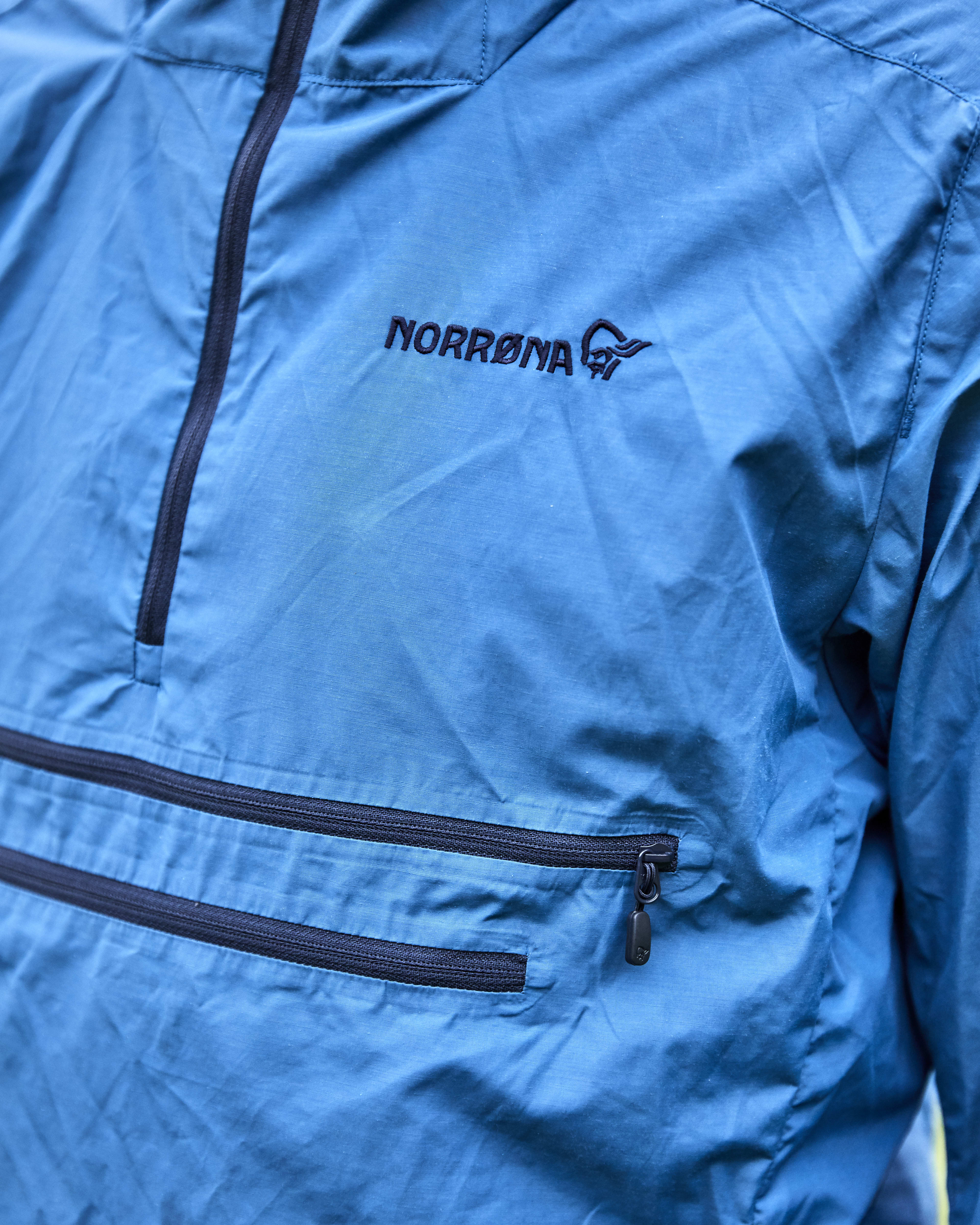 Closeup of a blue Norrøna jacket with a zipper and a pocket. 
