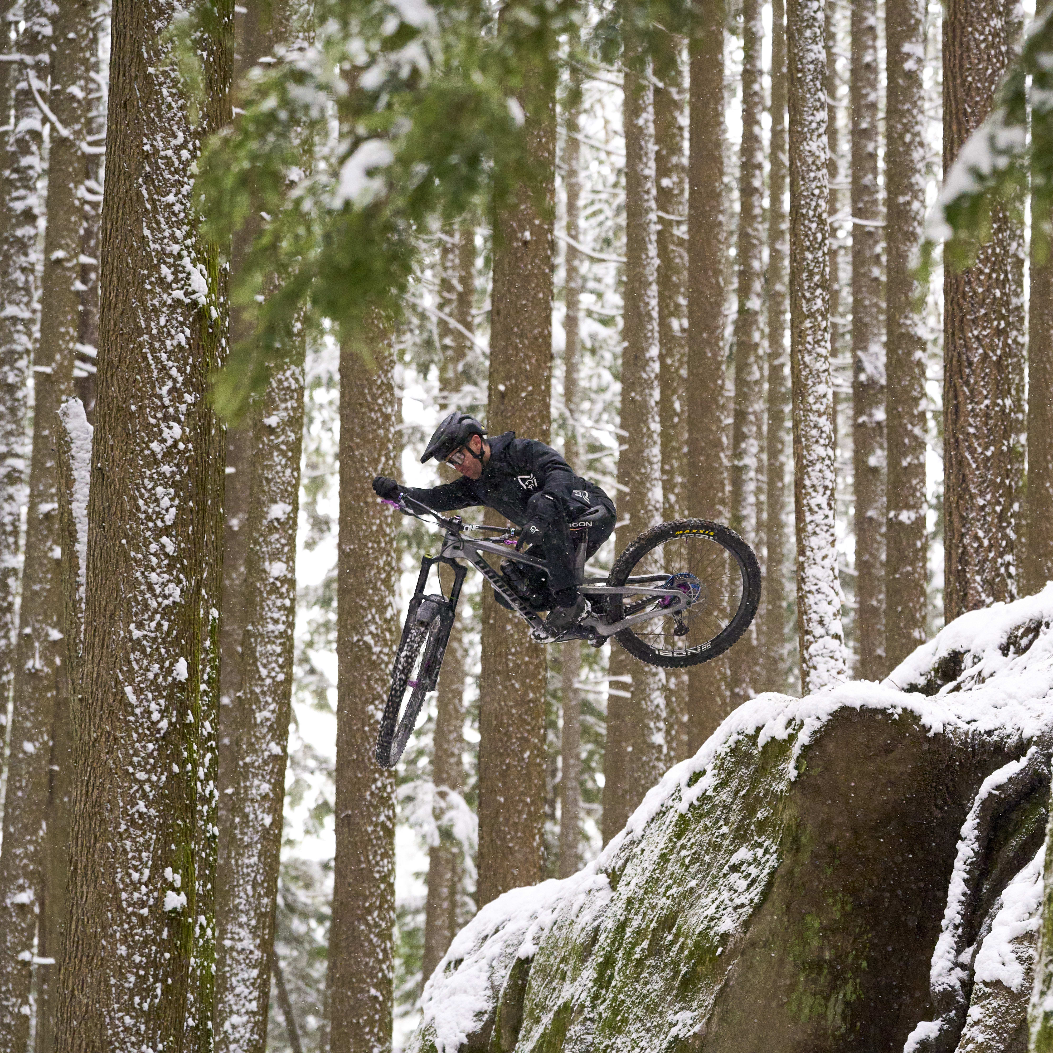 A person in Norrøna clothing riding a mountain bike over a snowy rock in a forest.