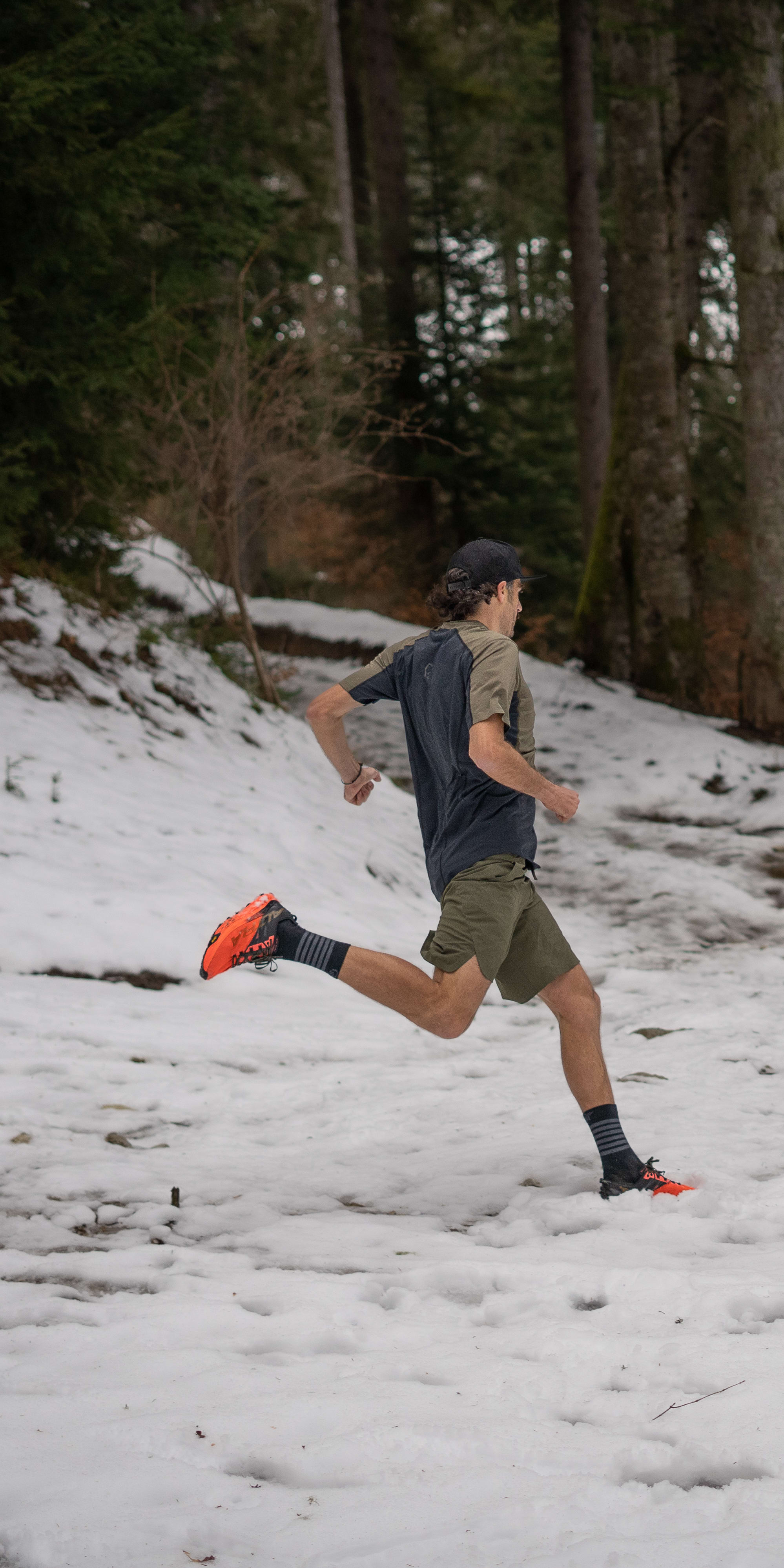 A man wearing a Norrøna shirt and shorts runs through a snowy forest.