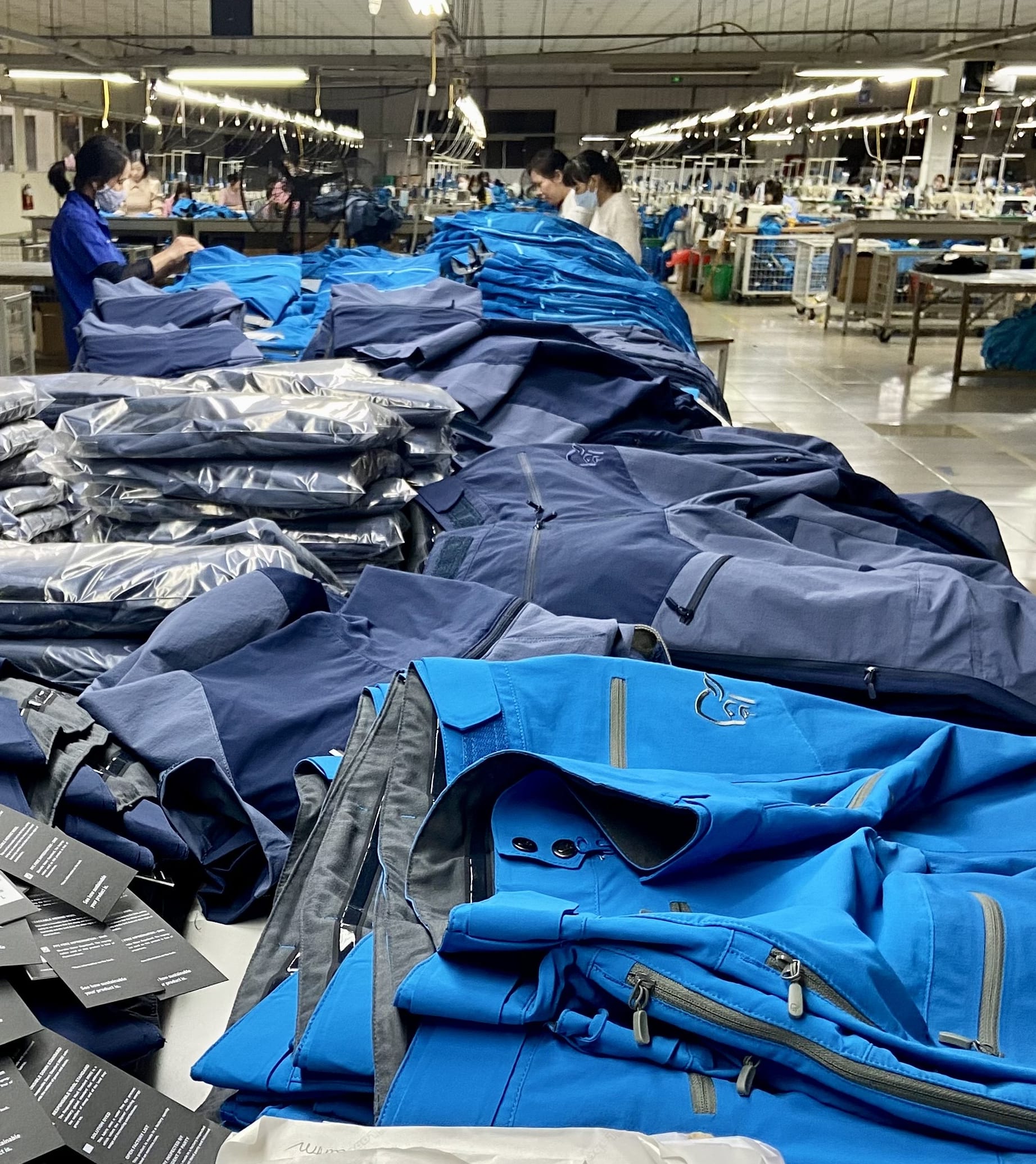 A stack of folded Norrøna clothing, mostly in blue and black, with several tags visible. A worker stands near the pile in a factory setting.
