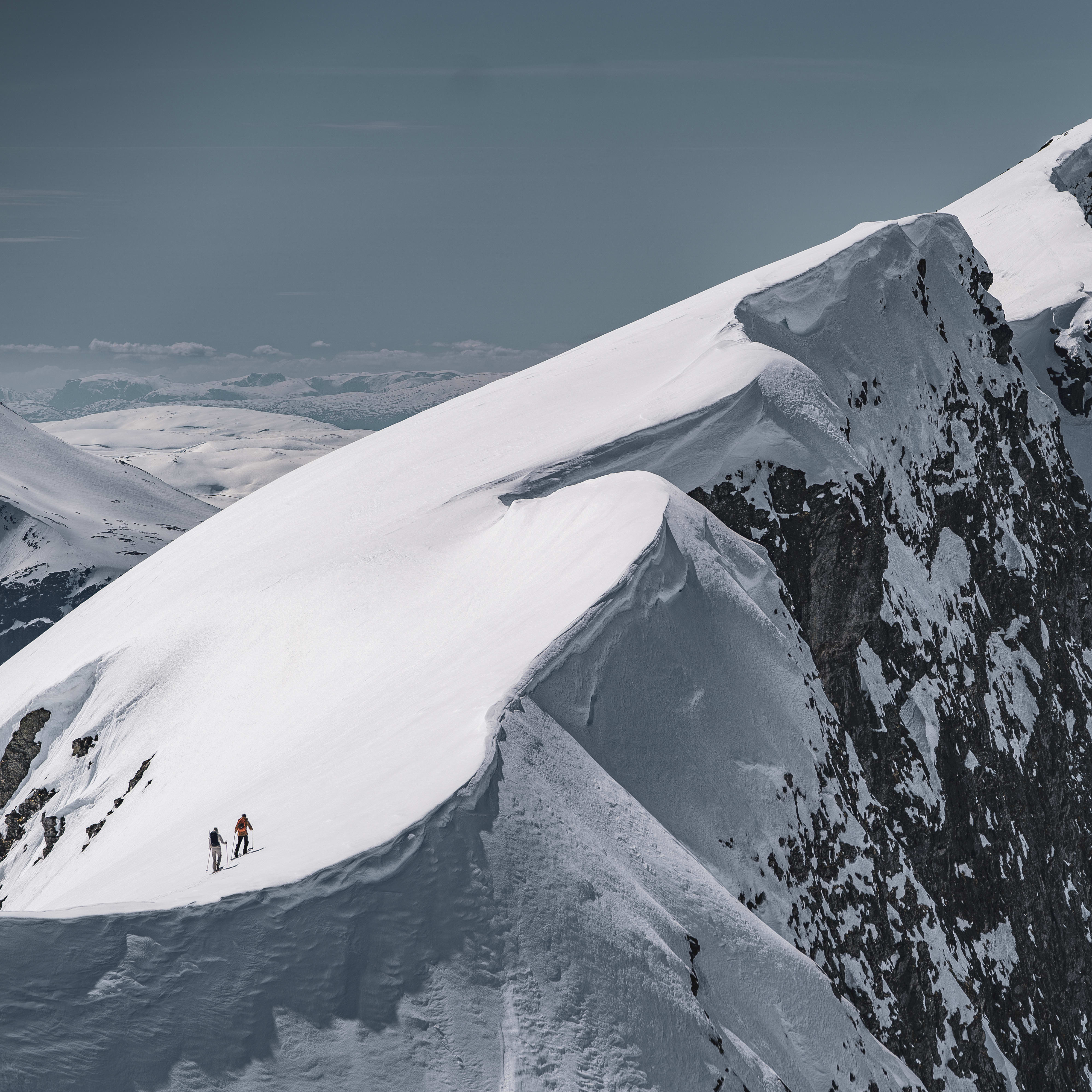 Two people in Norrøna gear hike along a snowy mountain ridge with a steep rocky cliff on the right.  
