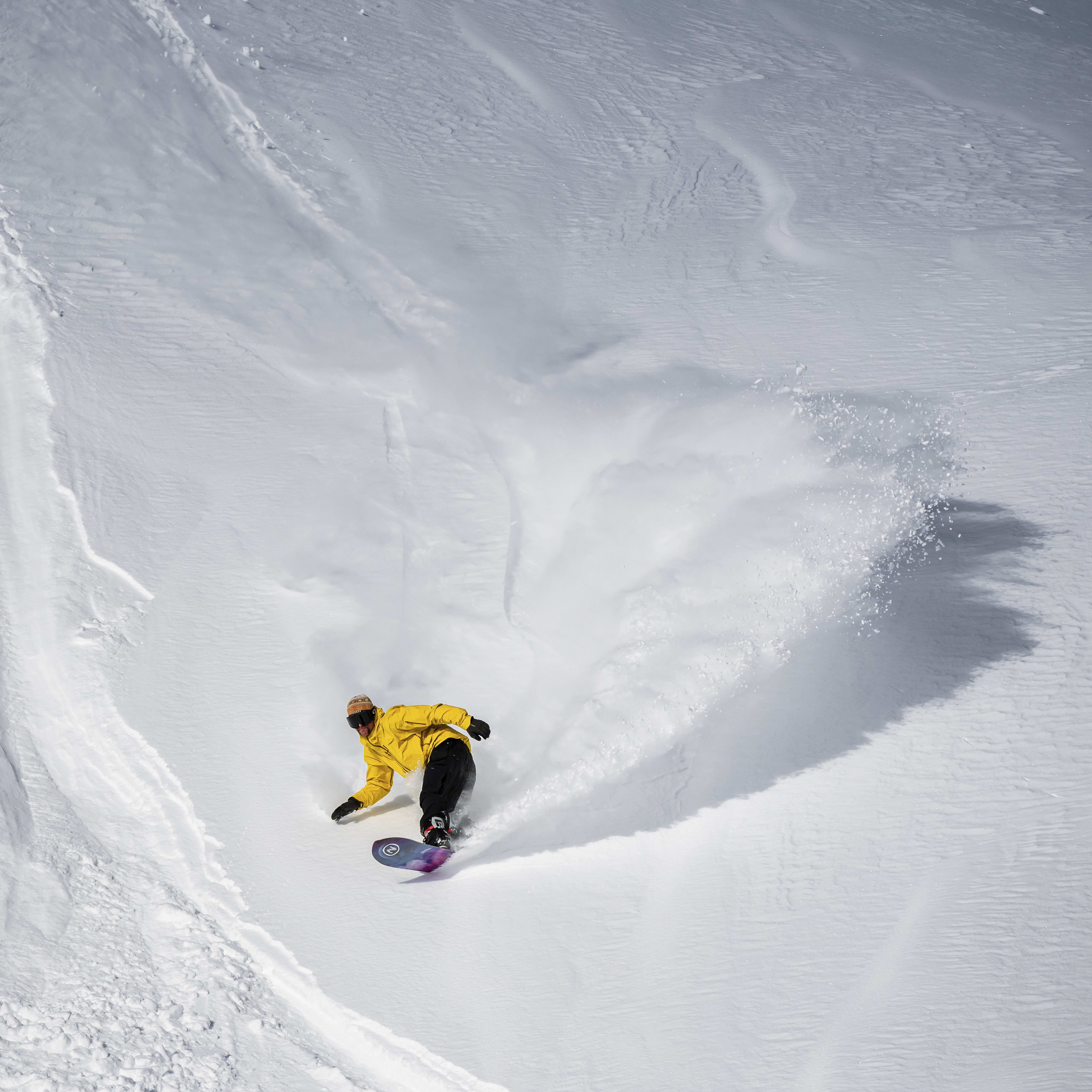 A snowboarder in a yellow jacket and black pants rides down a snowy slope, leaving a trail of snow behind them. The snowboarder is wearing a hat and goggles.