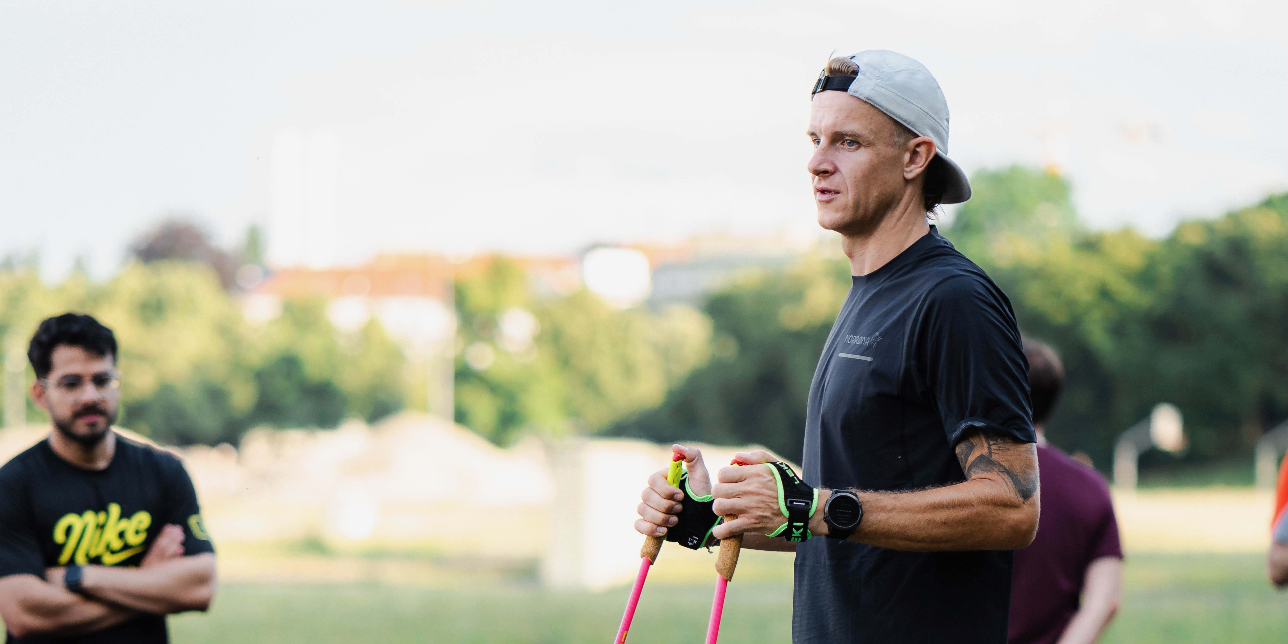 A man stands holding hiking poles, wearing a Norrøna T-shirt, shorts, cap, and a watch. Other people are in the background.