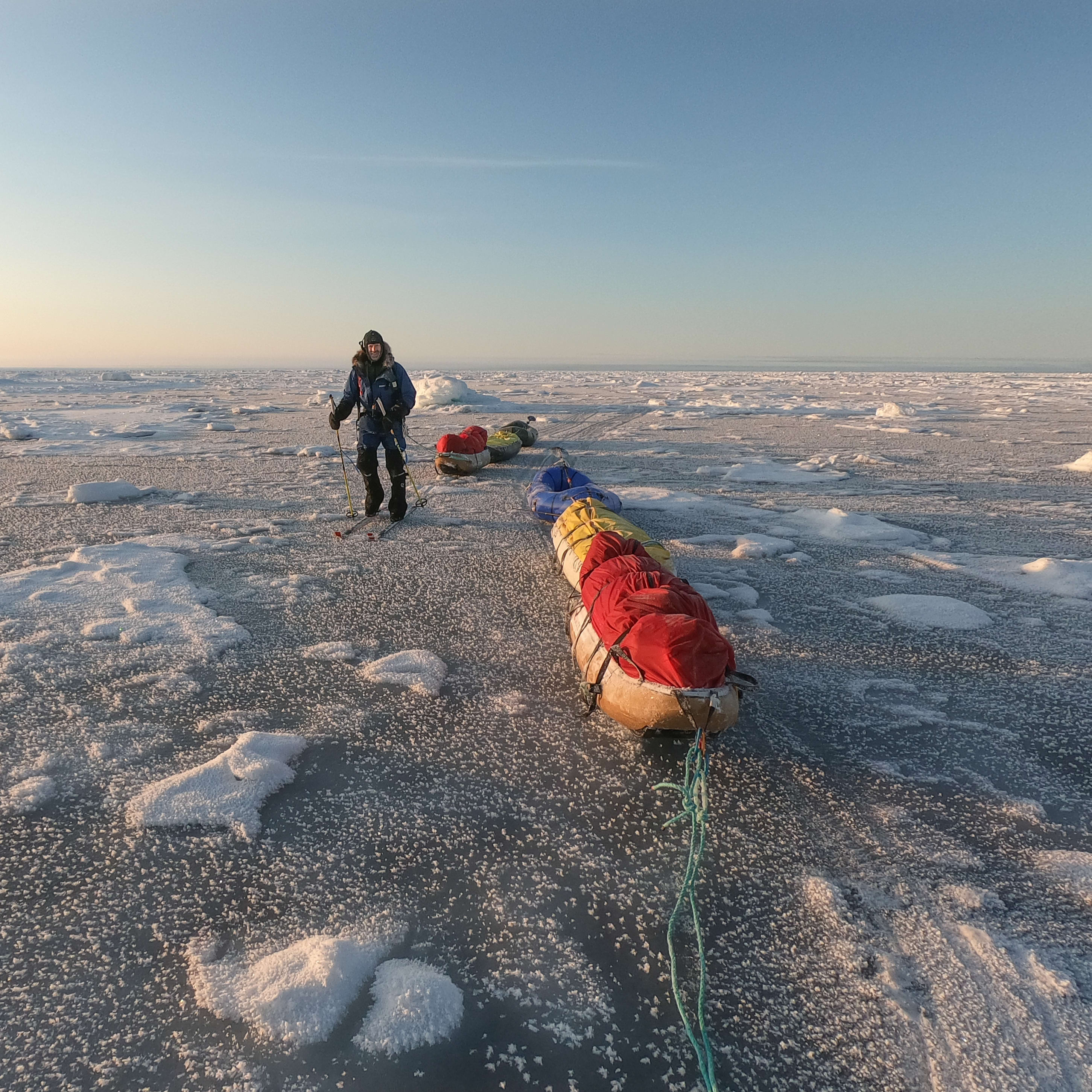A person wearing a Norrøna jacket cross-country skis across a frozen lake with a sled towed behind. 
