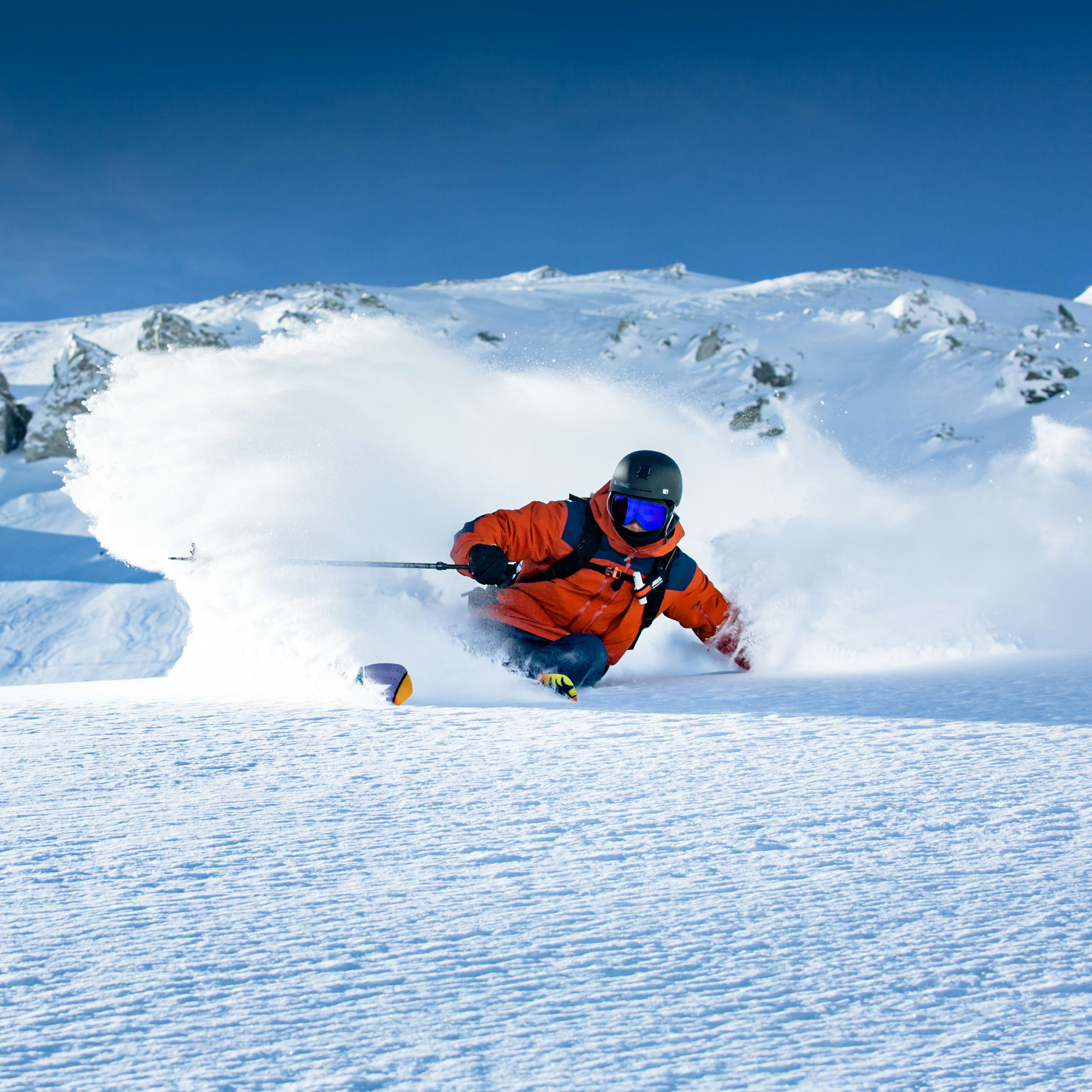 A skier wearing Norrøna gear makes a turn, kicking up a spray of snow.  The skier is wearing a helmet and goggles.  The sky is blue and the mountain is snow covered. 
