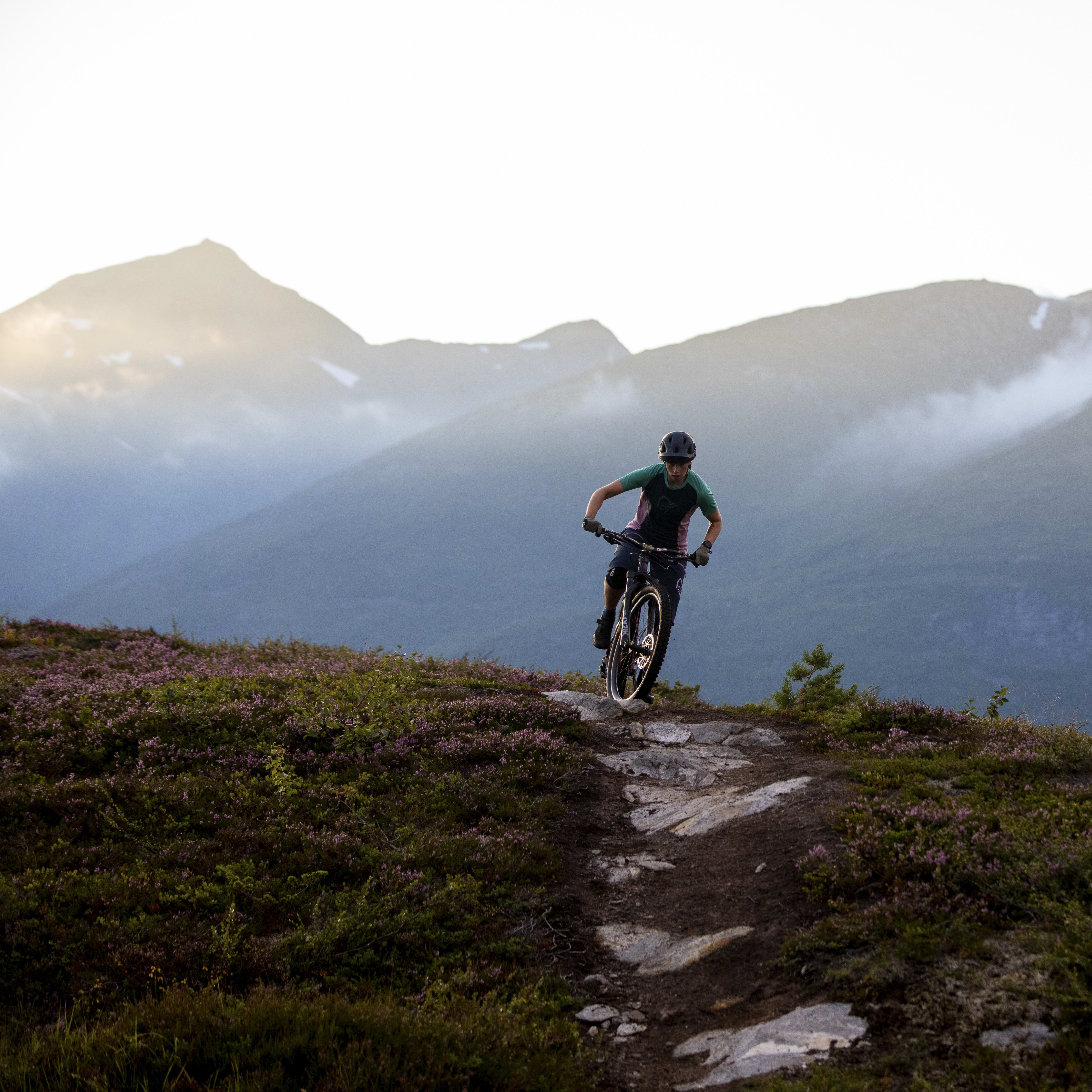 A person mountain biking wearing a helmet and a green Norrøna shirt on a trail with purple heather. Mountains are visible in the background.