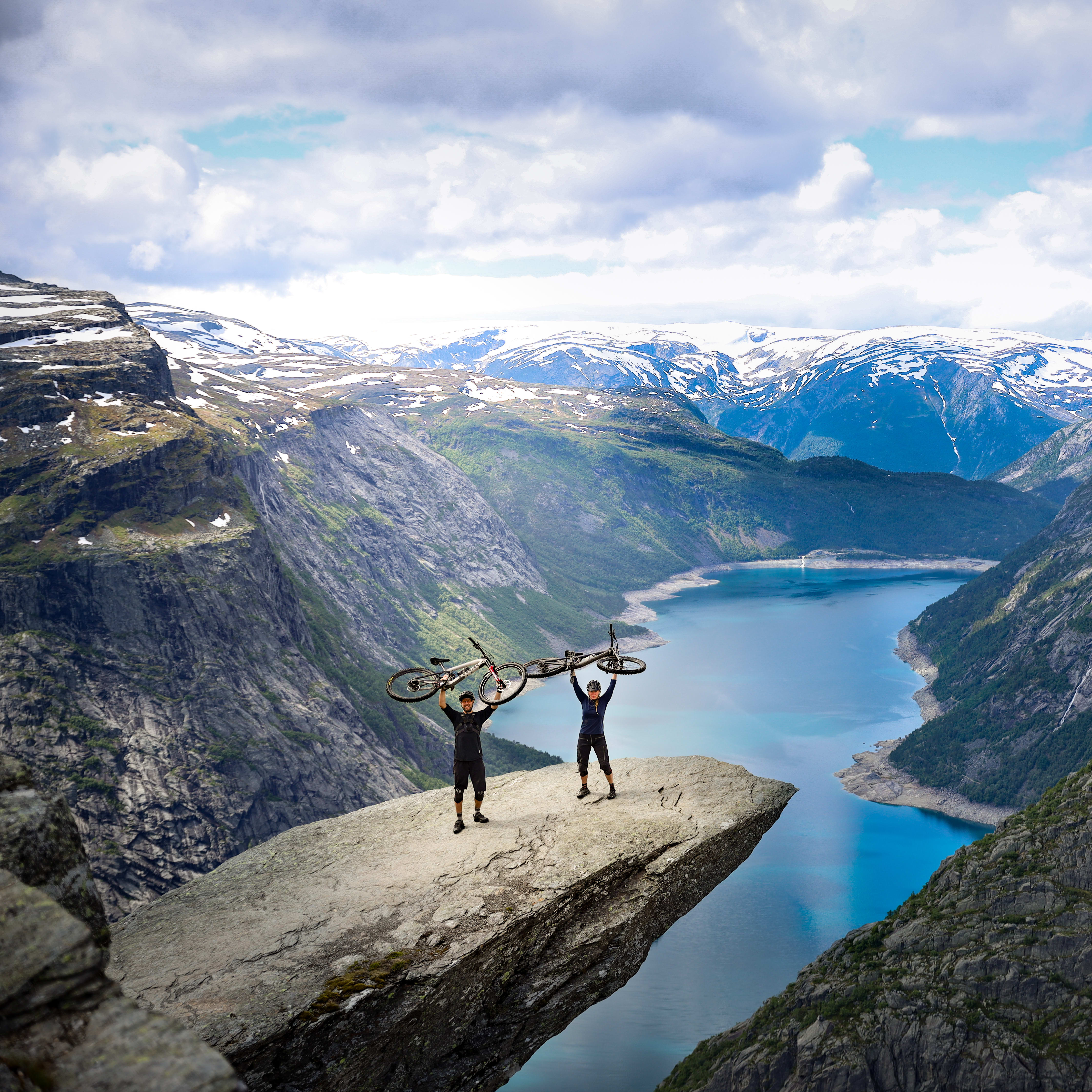 Two people stand on a cliff overlooking a fjord. They are holding up Norrøna mountain bikes.  
