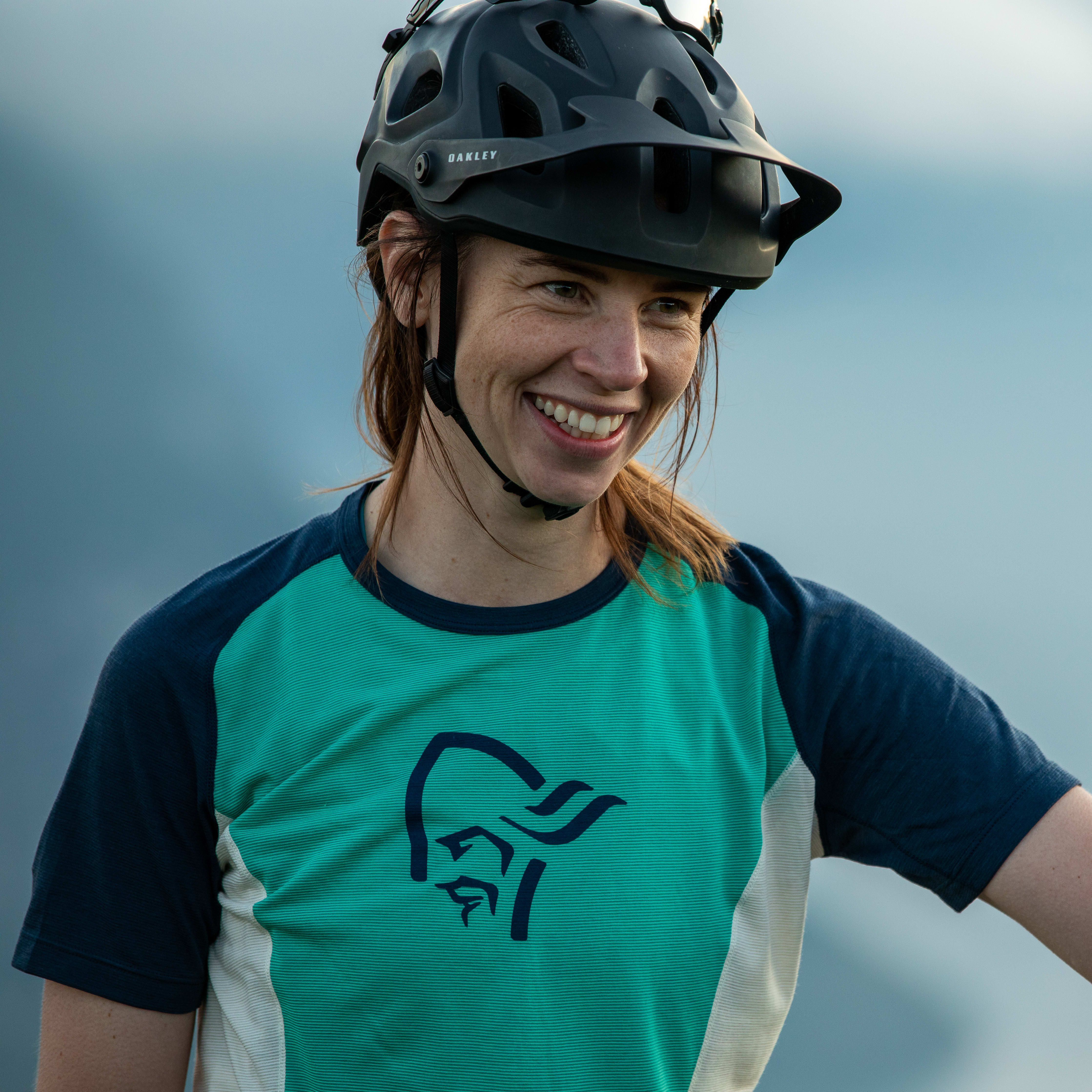 A woman wearing a Norrøna shirt and helmet smiles against a blue sky. 
