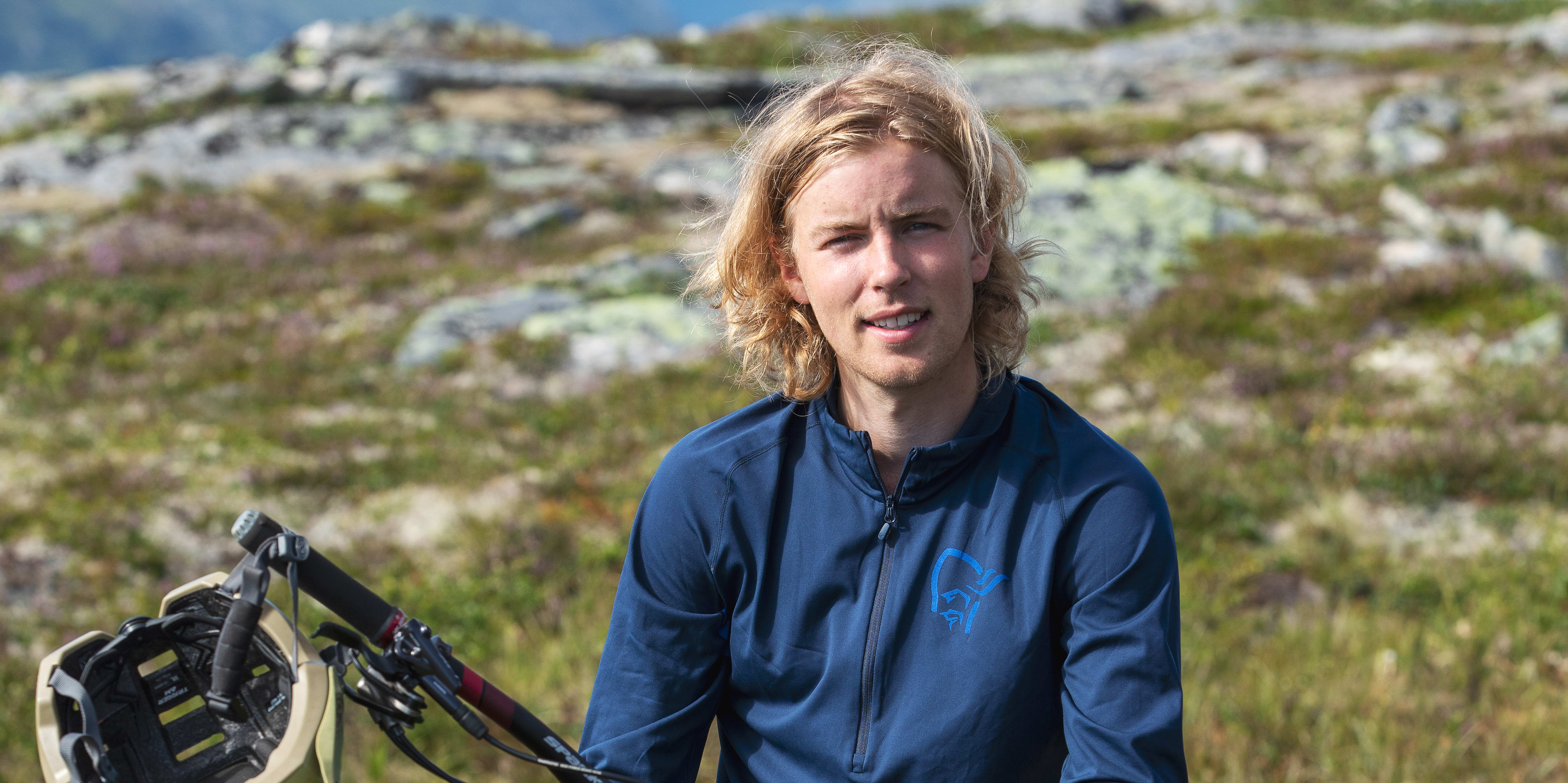 A man wearing a Norrøna shirt and shorts sits on a mountain bike in a grassy field.