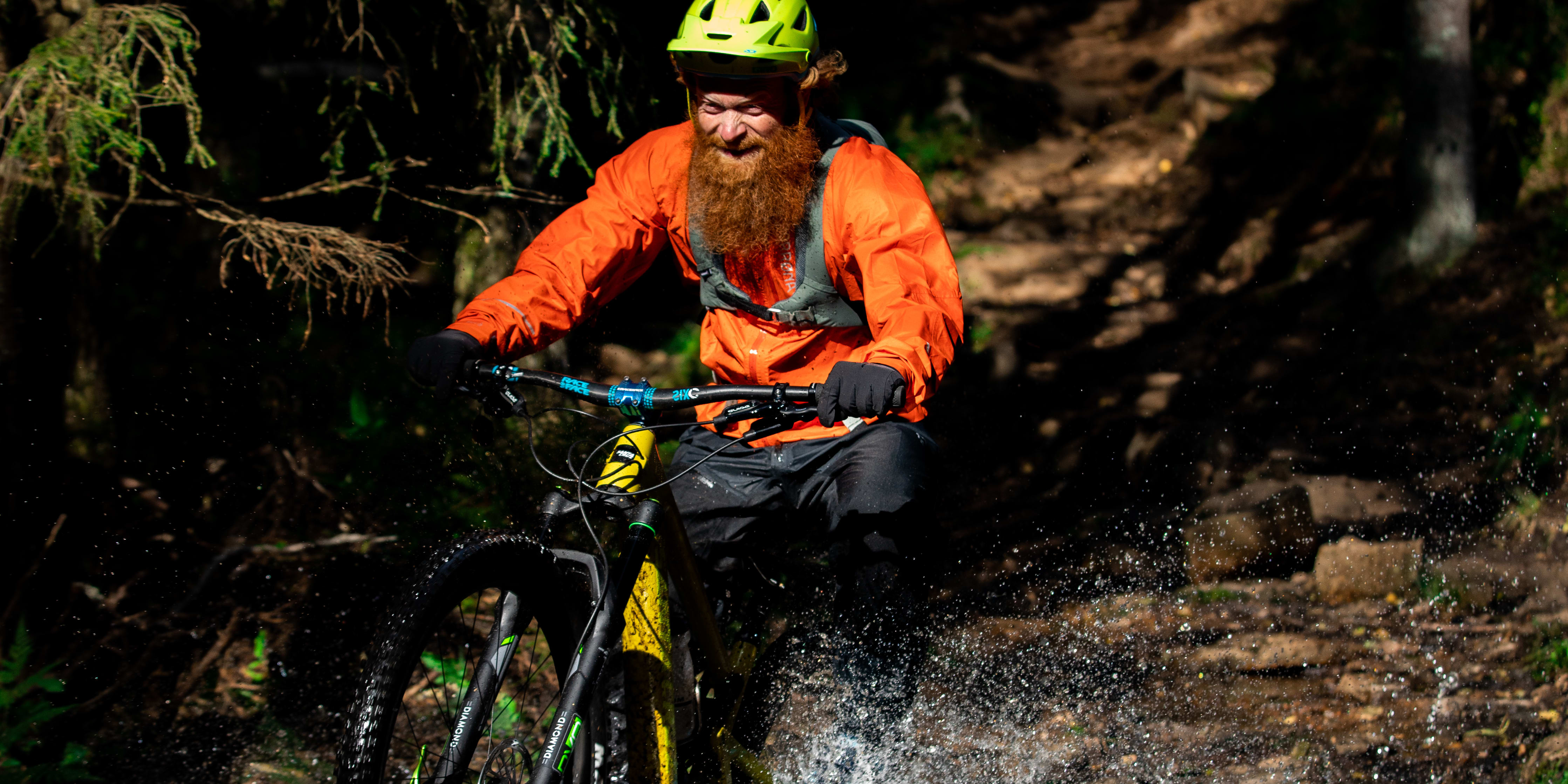 A man wearing a Norrøna jacket and a helmet rides a mountain bike through a forest.  Water splashes up from his tires as he goes. 
