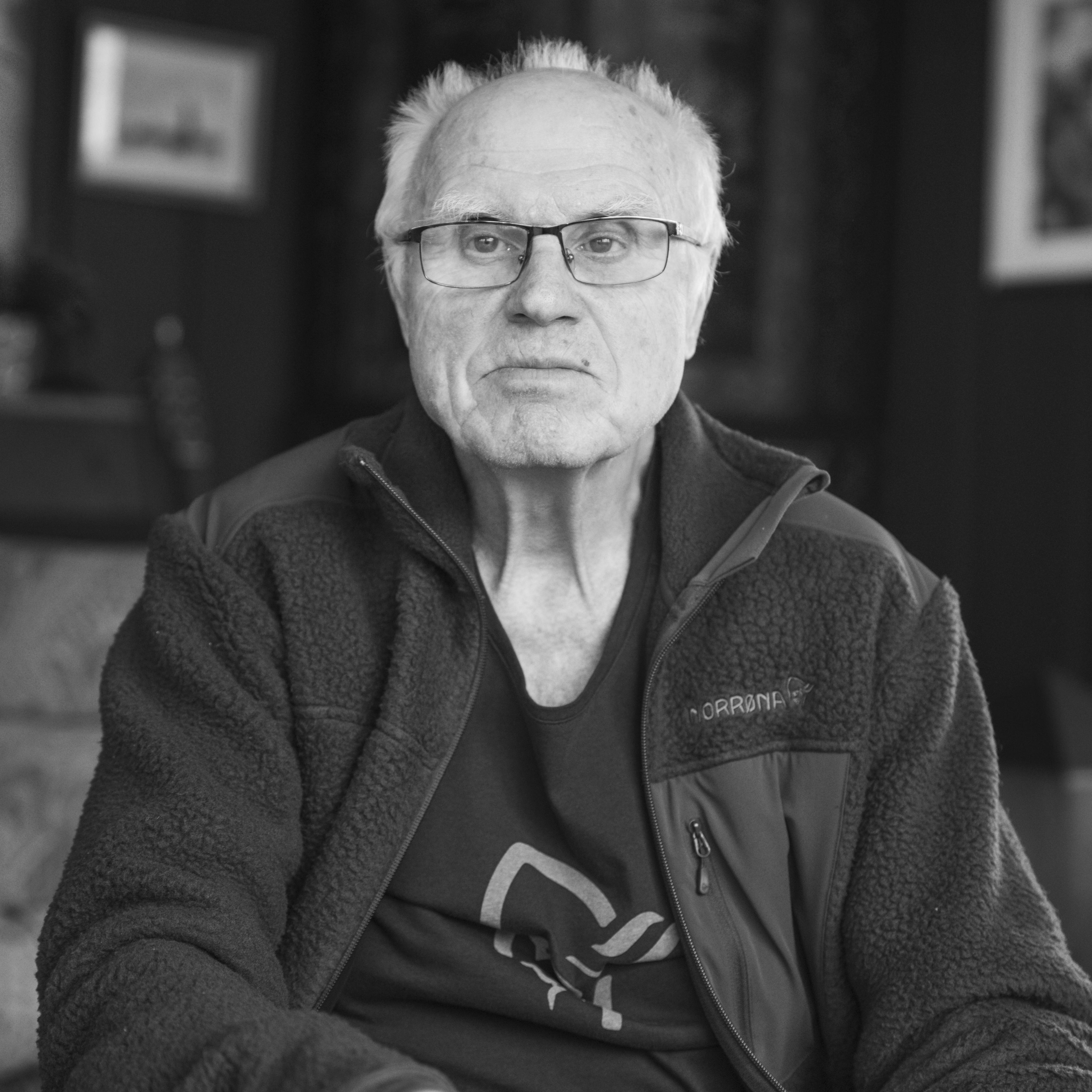 Black and white photo of a man wearing a Norrøna jacket. He has glasses and short white hair. He is looking directly at the camera.