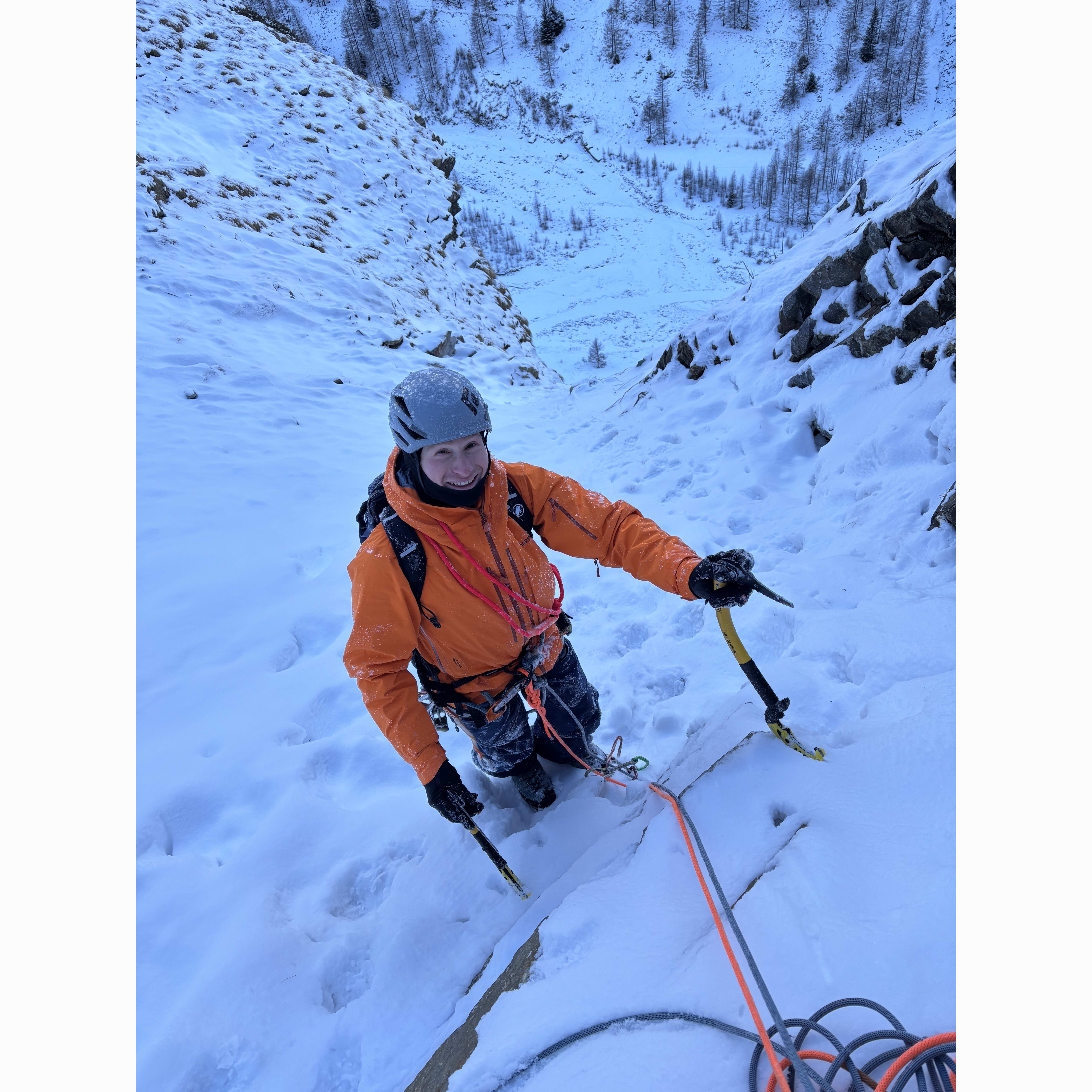 A smiling ice climber wearing a helmet and Norrøna gear ascending a snowy ice wall.