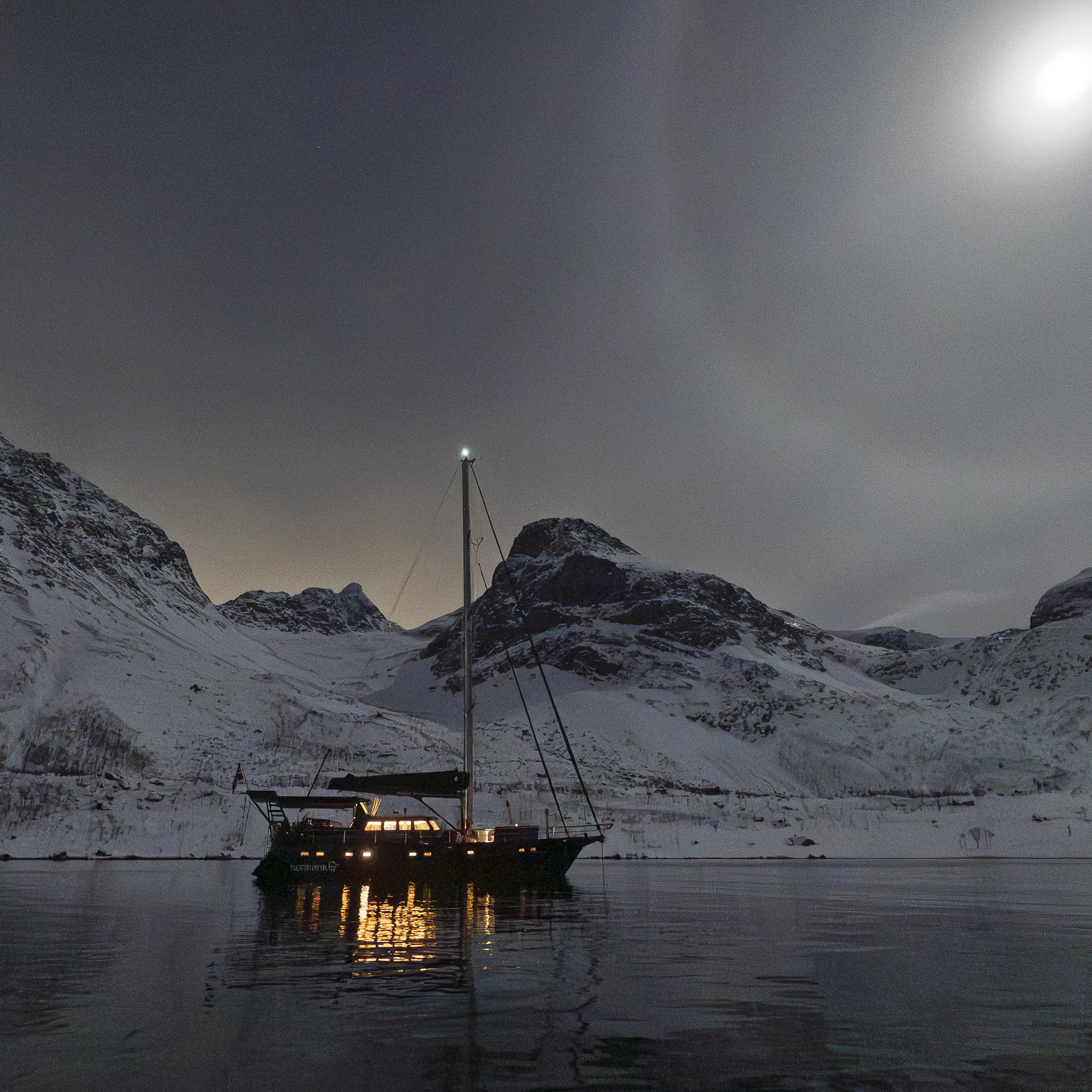 Moonlit view of a sailboat on dark water, with snow-covered mountains in the background.