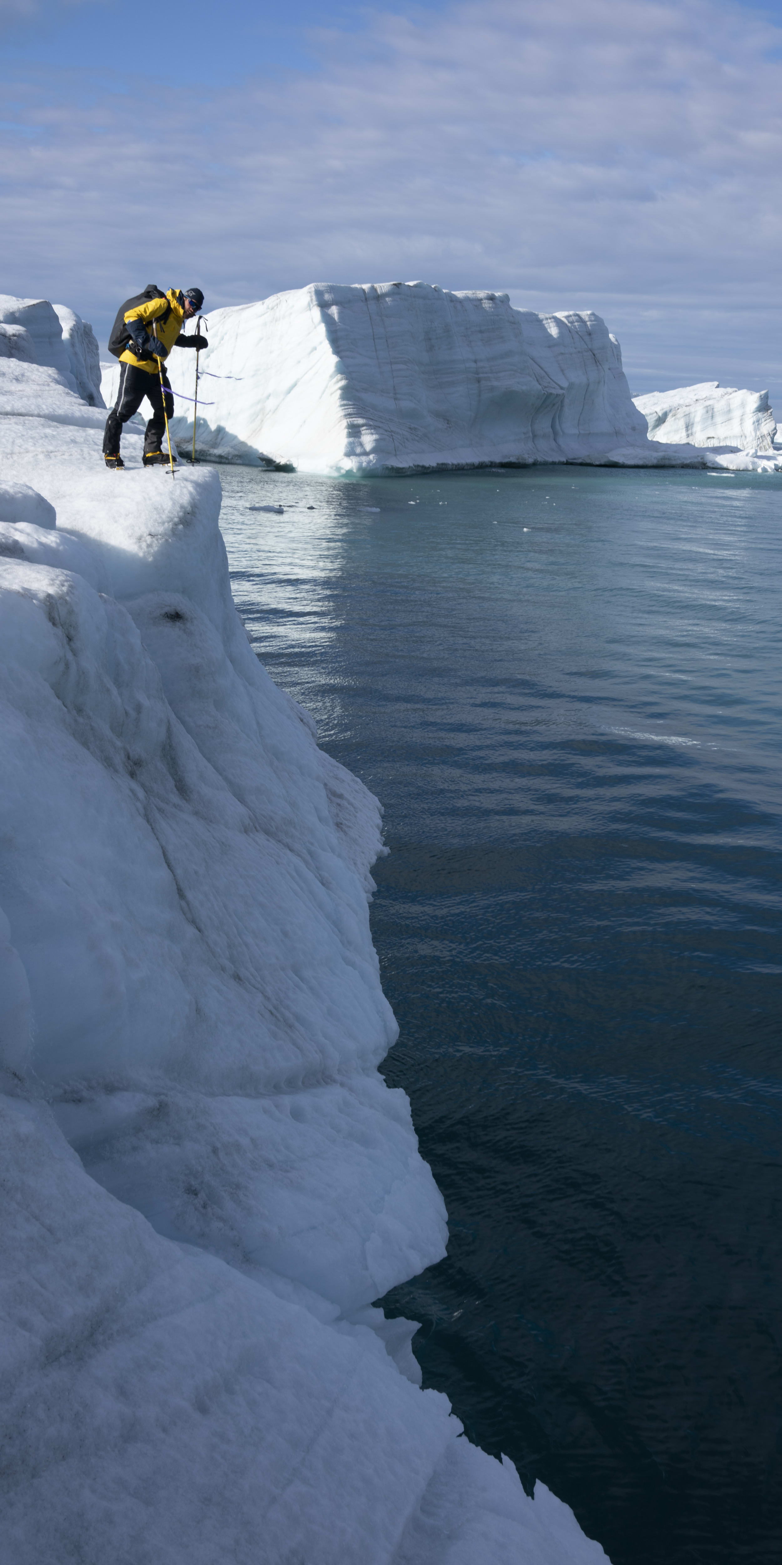A person is standing on the edge of an ice cliff, overlooking a blue body of water.  The person is wearing a yellow jacket and is holding ice climbing equipment.  In the background, there is a large ice berg.