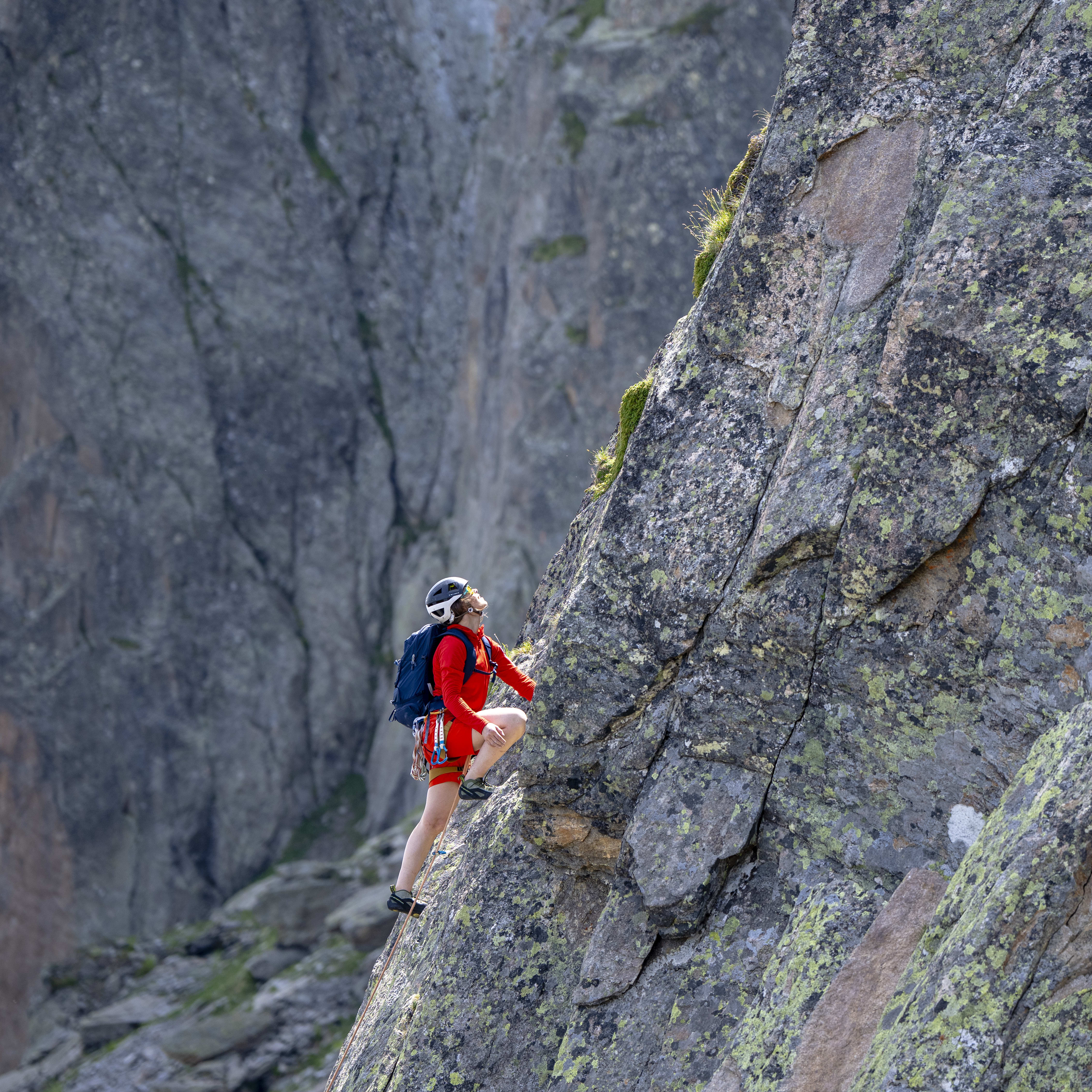 A rock climber in red Norrøna gear ascends a steep, lichen-covered rock face. The person wears a helmet and backpack with dramatic mountain backdrop.