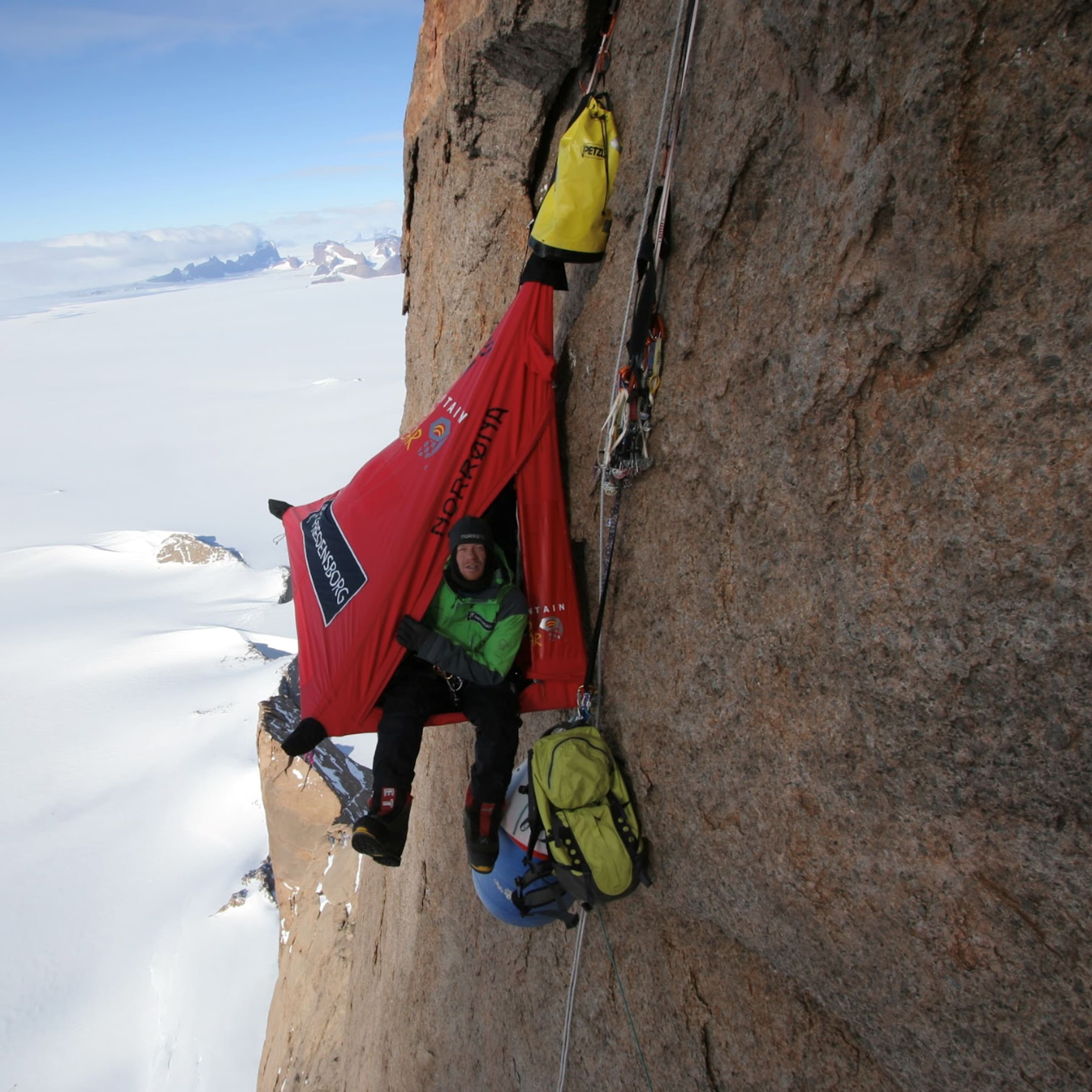 A climber is sitting in a red Norrøna tent hanging from a cliff, with a vast snowy landscape below.