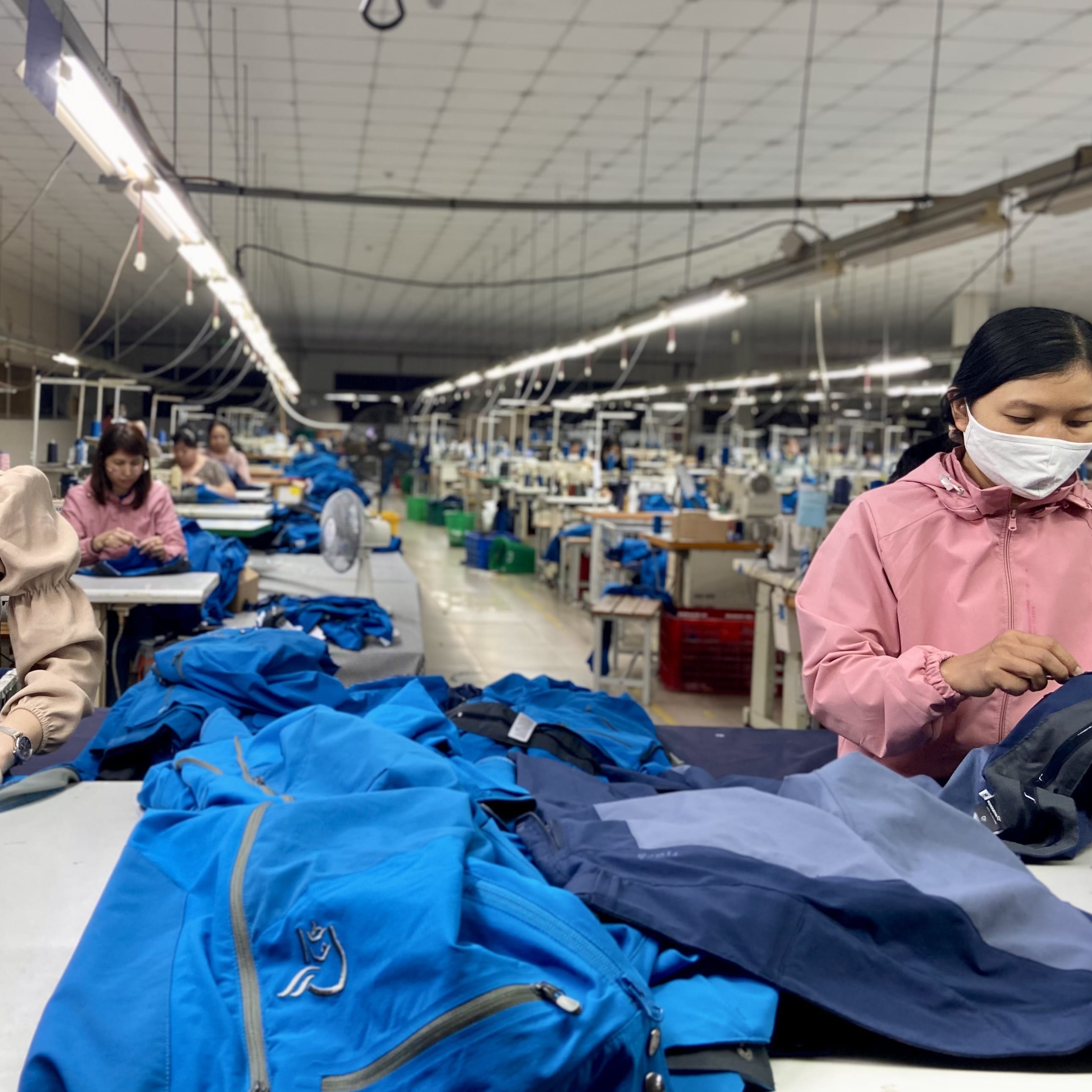 Two women working in a factory sewing Norrøna clothing.  A stack of blue jackets with the Norrøna logo is in the foreground. 
