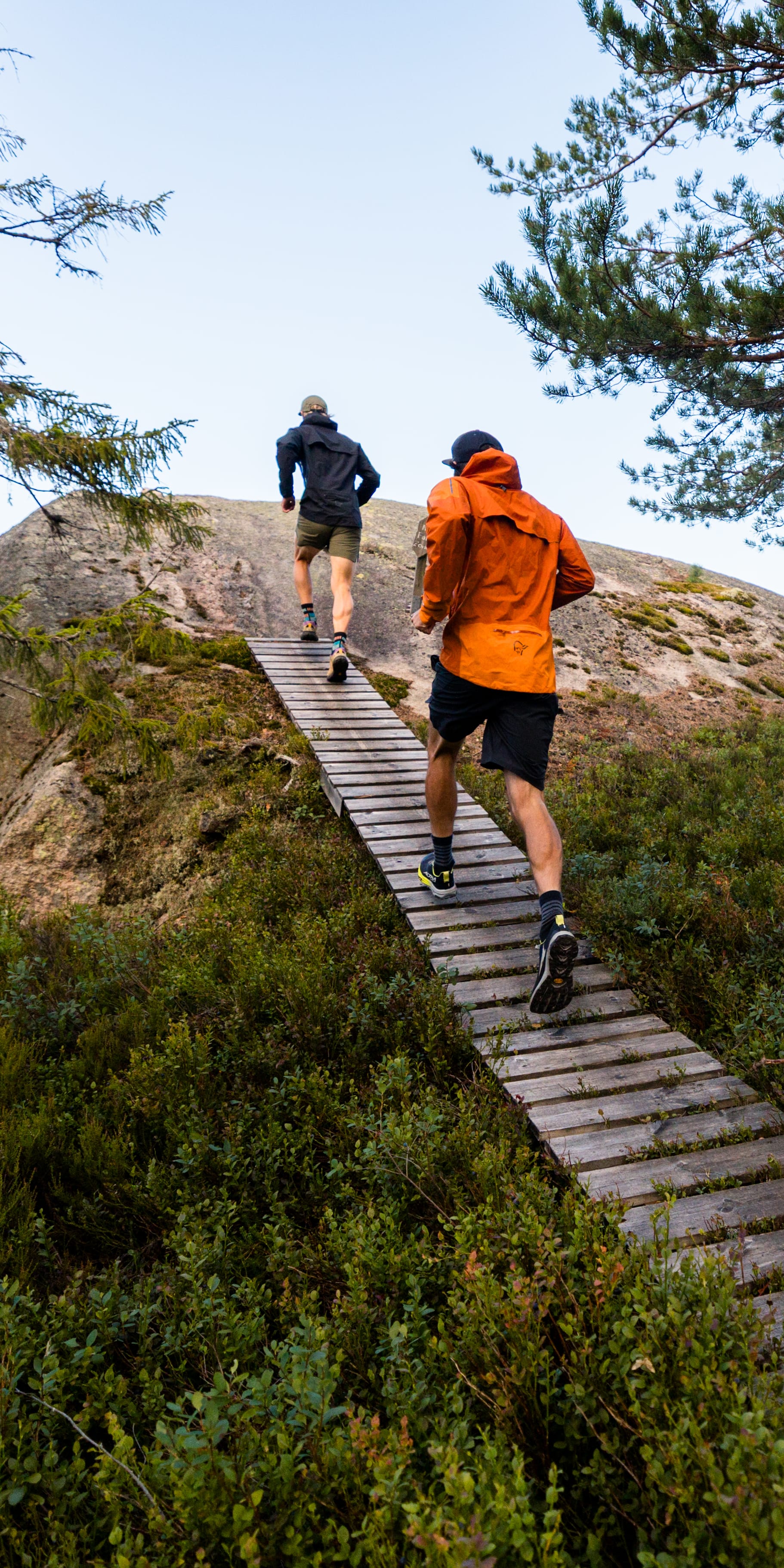 Two runners on a wooden trail in a forest. The runner in front wears an orange Norrøna jacket.
