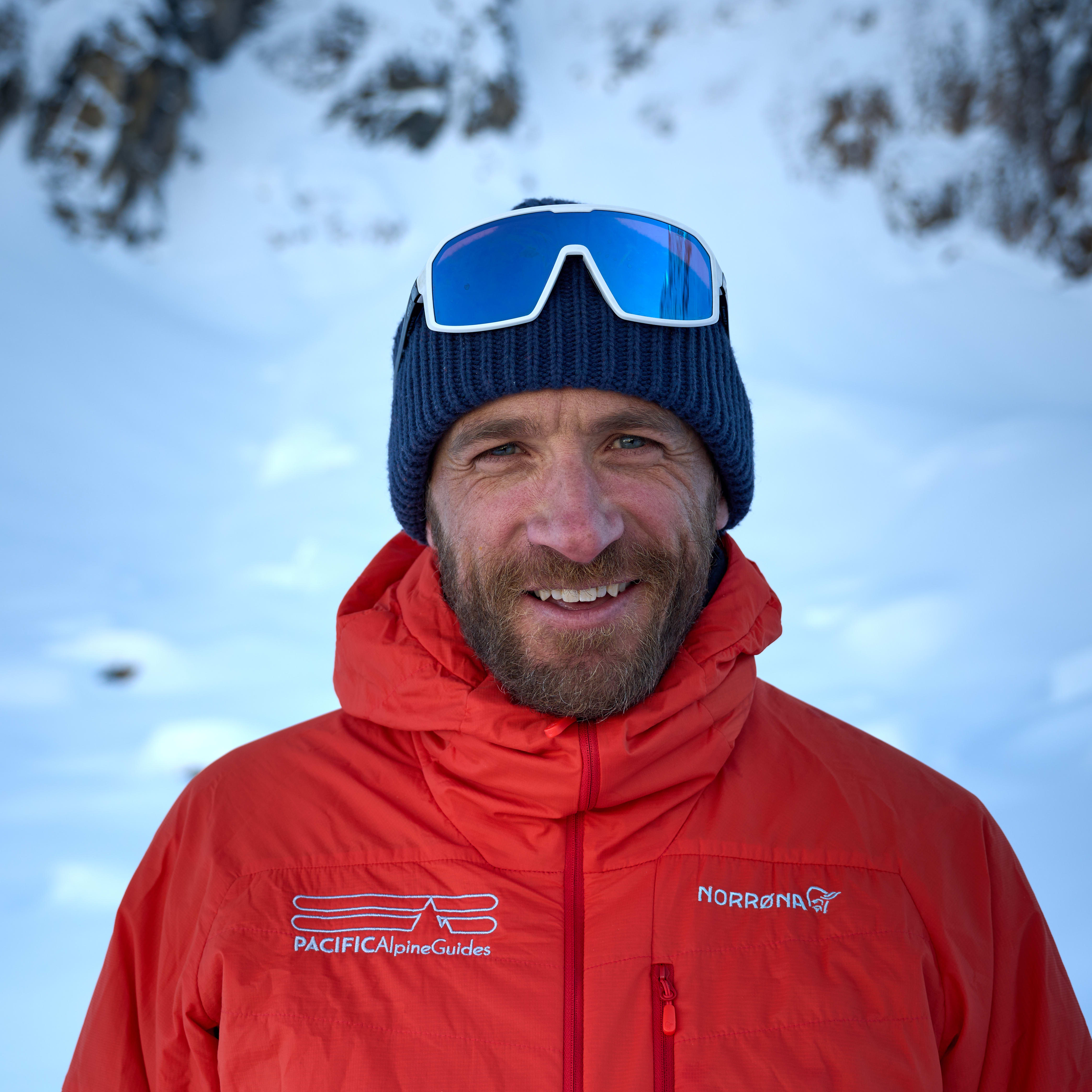 A smiling man with a beard, wearing a blue beanie, white goggles and a red Norrøna jacket. Snow in the background.