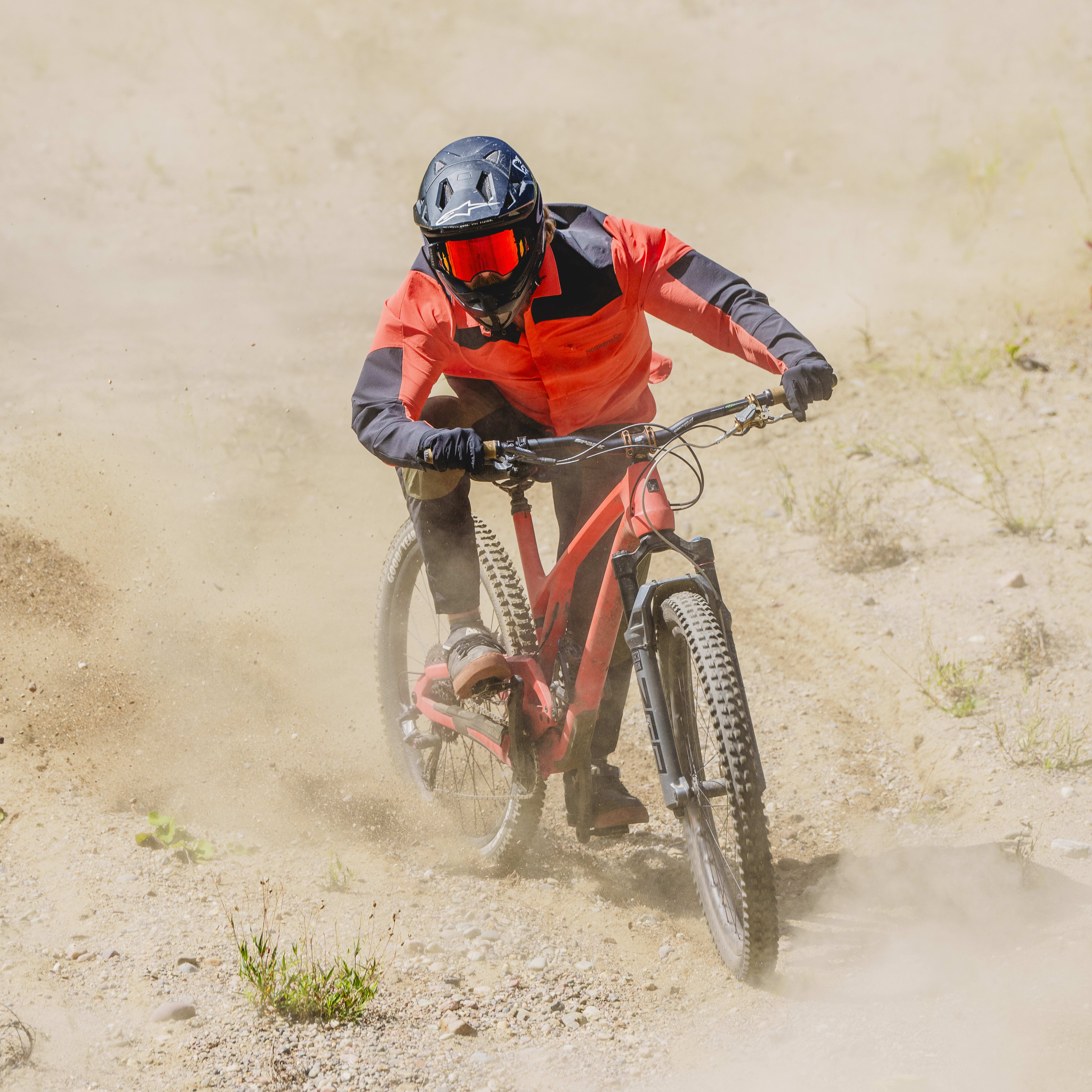 A person wearing a Norrøna jacket rides a mountain bike on a dirt path, kicking up dust. 
