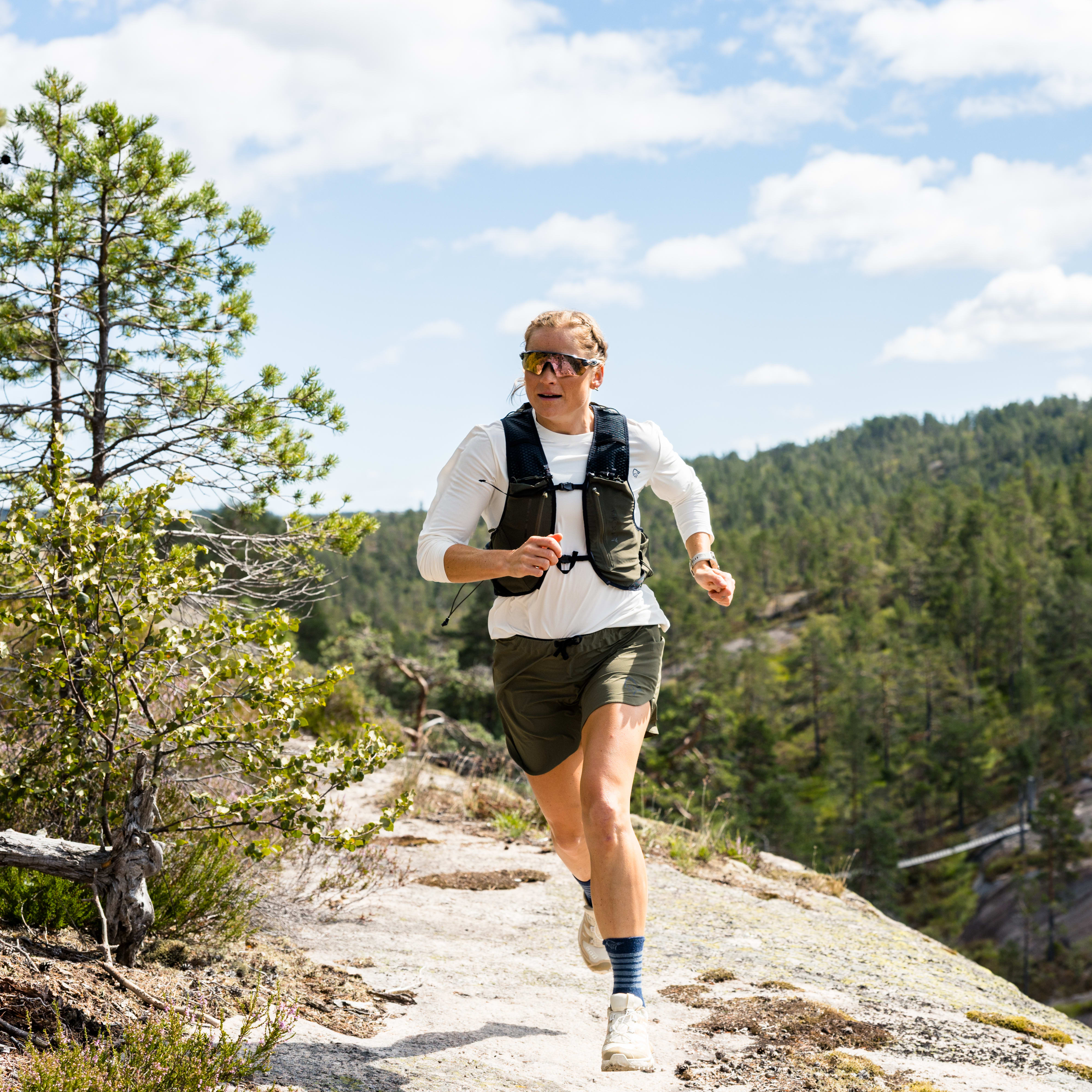 A person trail running in nature, wearing a Norrøna vest and shorts. The background shows trees and a partly cloudy sky.
