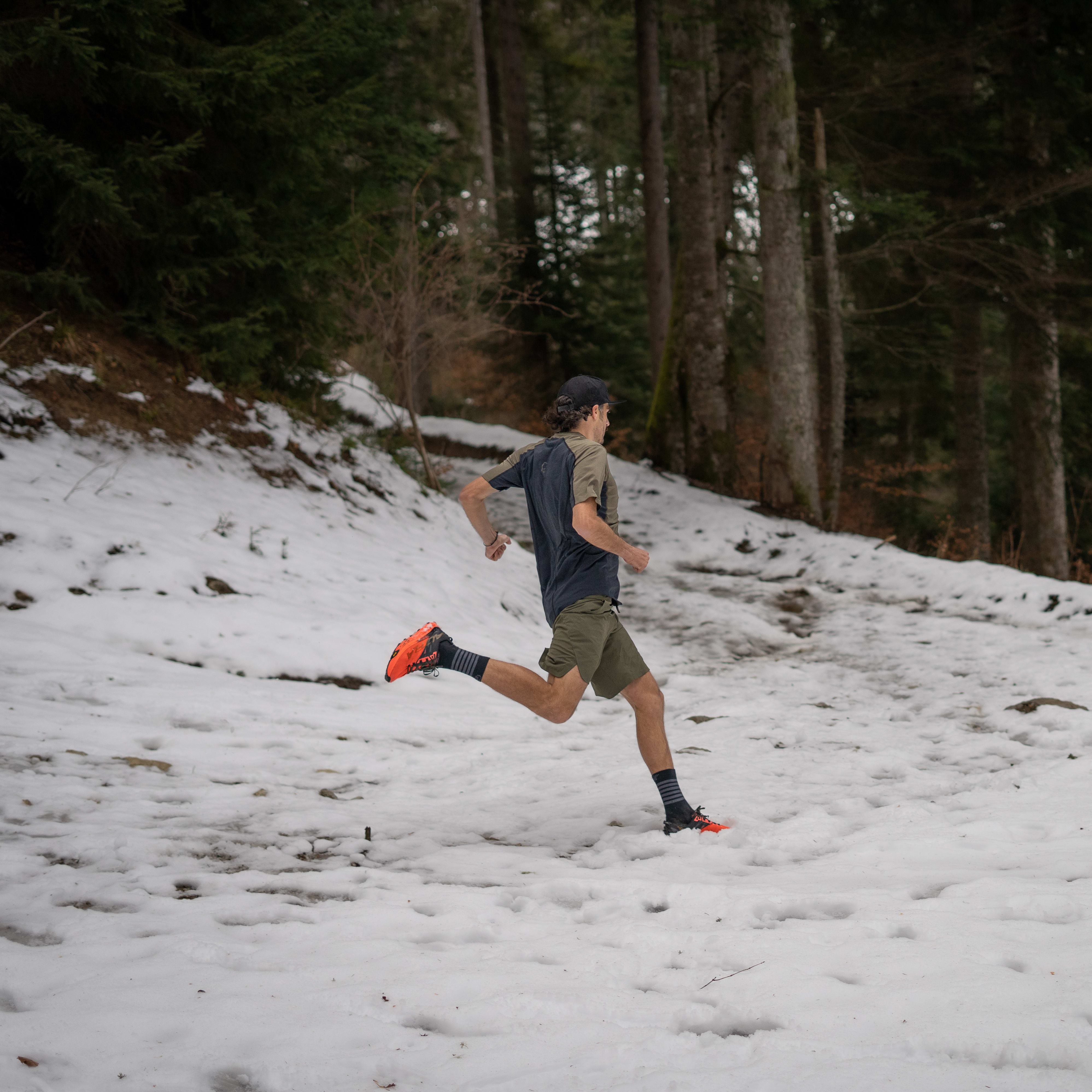 A man wearing a Norrøna shirt and shorts runs through a snowy forest. 
