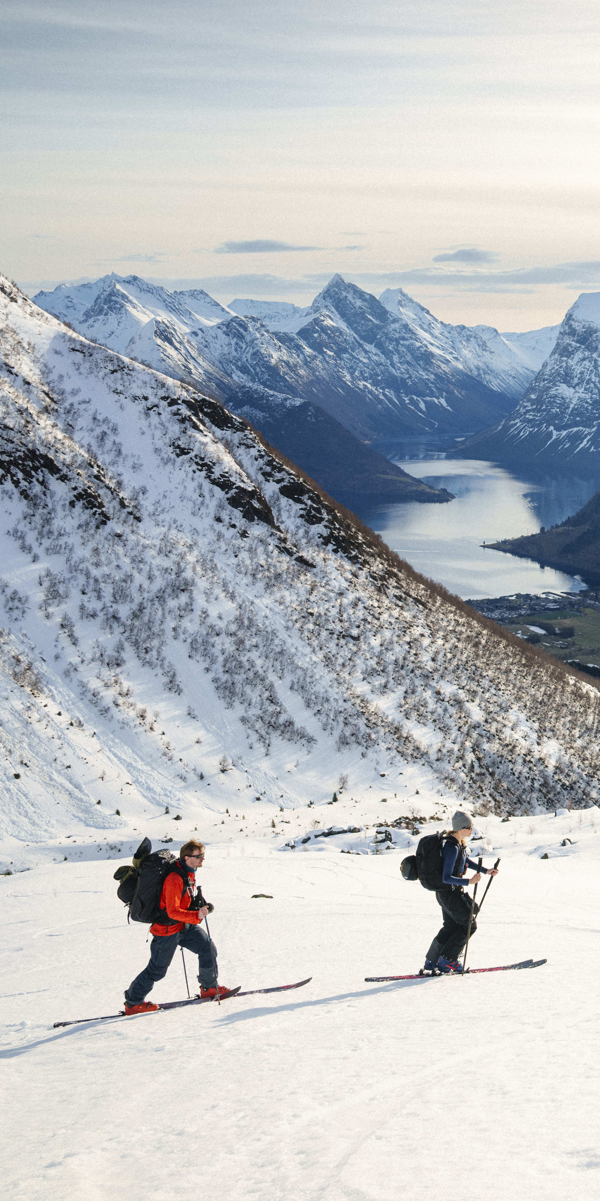 Four people ski down a snowy mountain with a fjord in the background. They are wearing Norrøna gear.  
