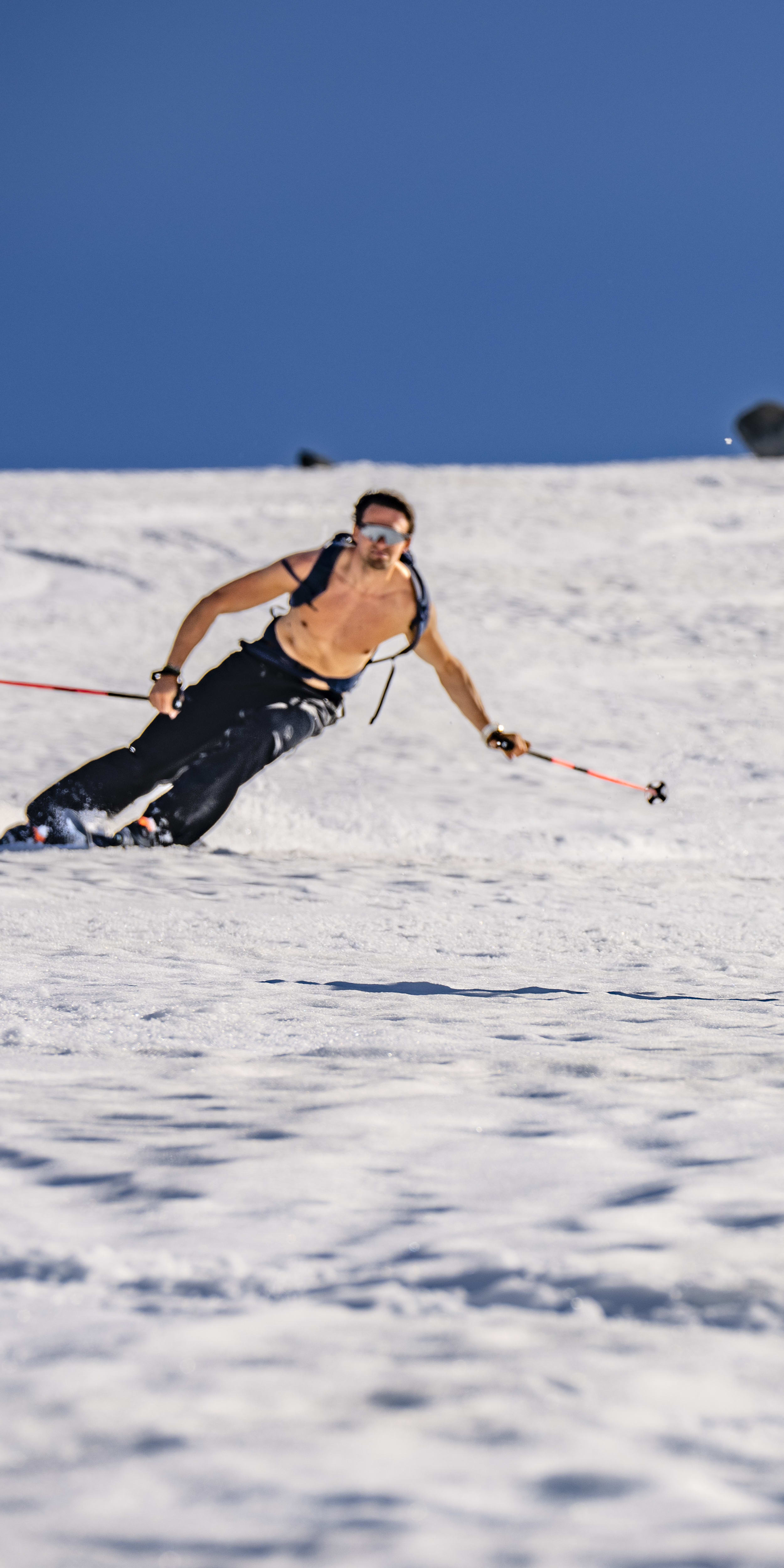 Two people ski down a snowy slope, a person in a blue plaid shirt is in the foreground, and a person in a black shirt is in the background. Both skiers are holding poles. The sky is blue.
