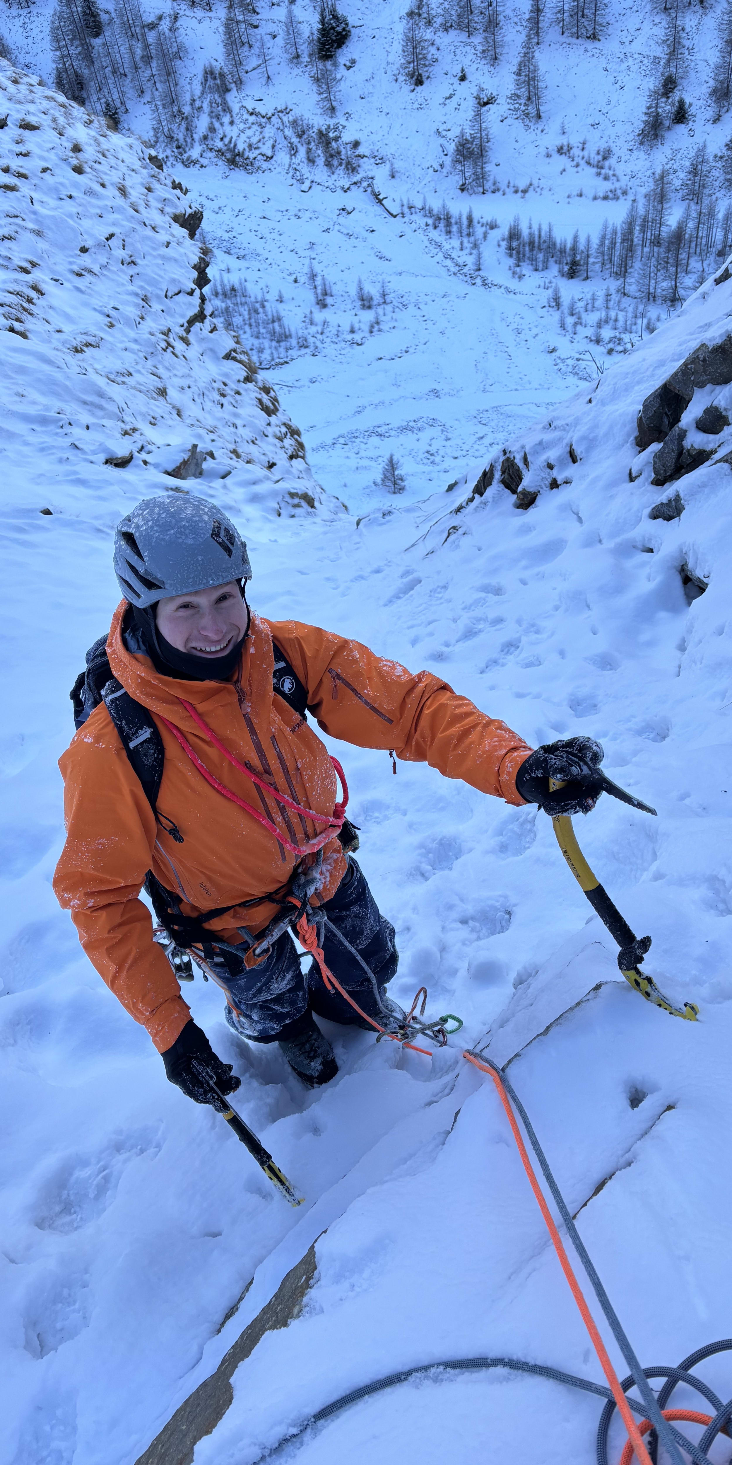 A smiling ice climber wearing a helmet and Norrøna gear ascending a snowy ice wall.  
